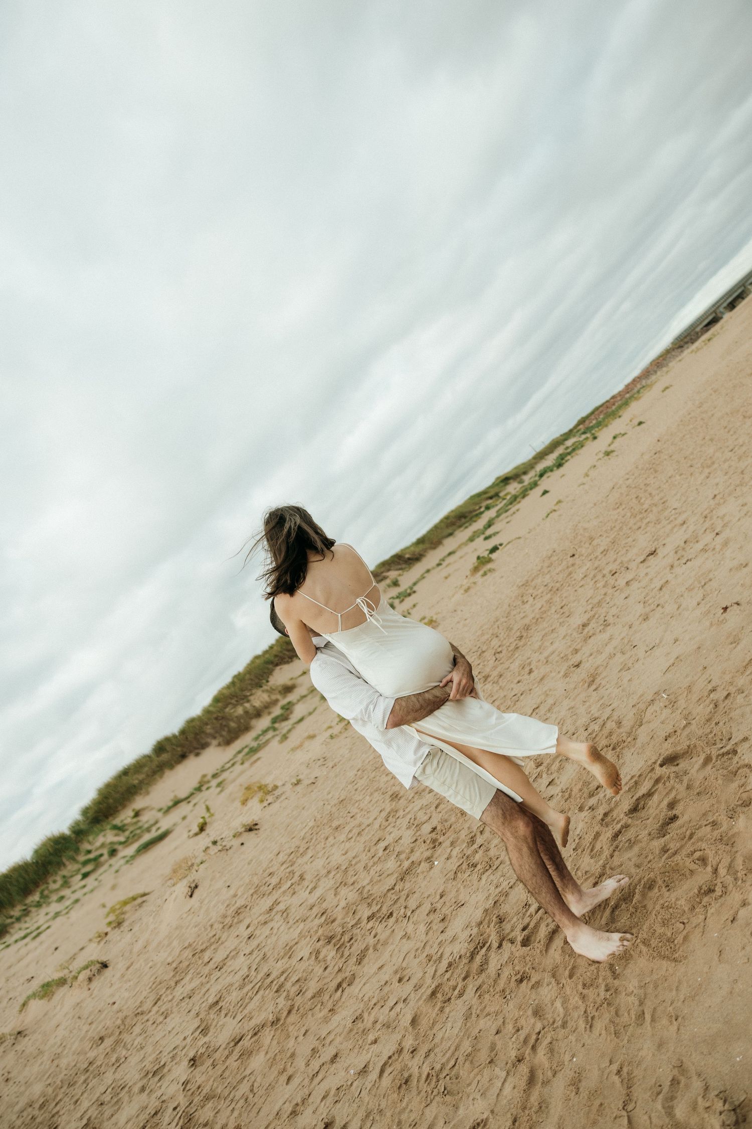 A person in white clothing lying artistically on a sandy beach with dramatic cloudy skies.