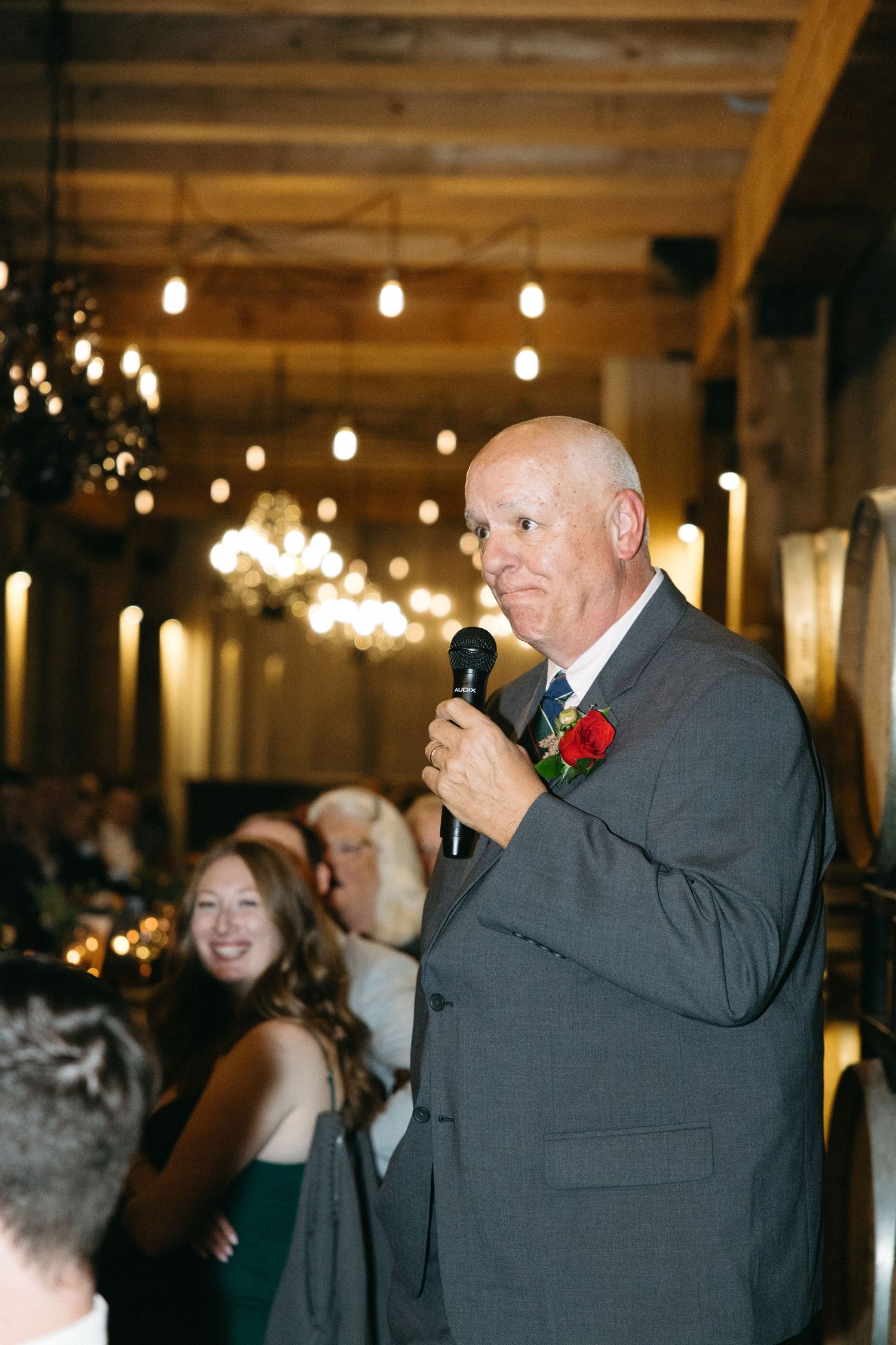 A speaker in a grey suit addresses guests at an event with string lights overhead.