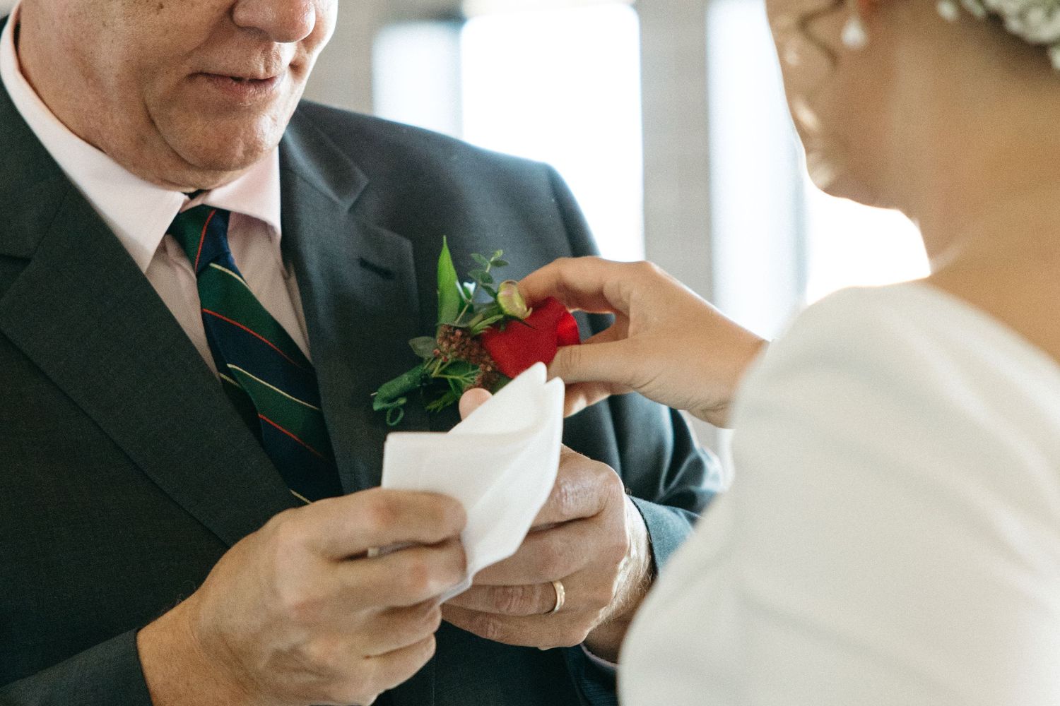 Close-up of a boutonniere being pinned onto a dark suit jacket during a wedding ceremony.