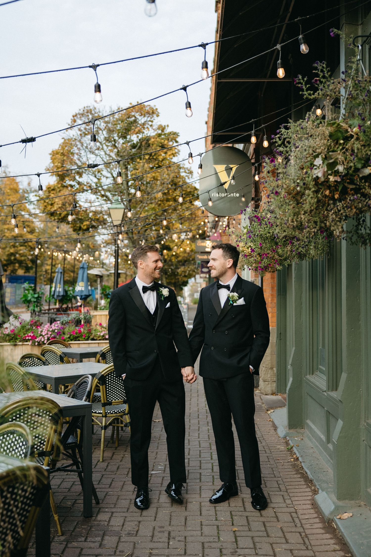 Wedding party members walk along a brick path with string lights and autumn trees overhead.