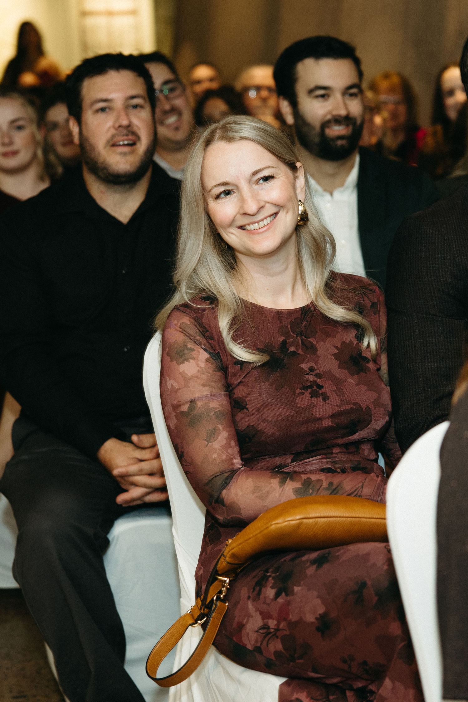 Audience members seated in a venue with someone wearing a burgundy dress in the foreground.