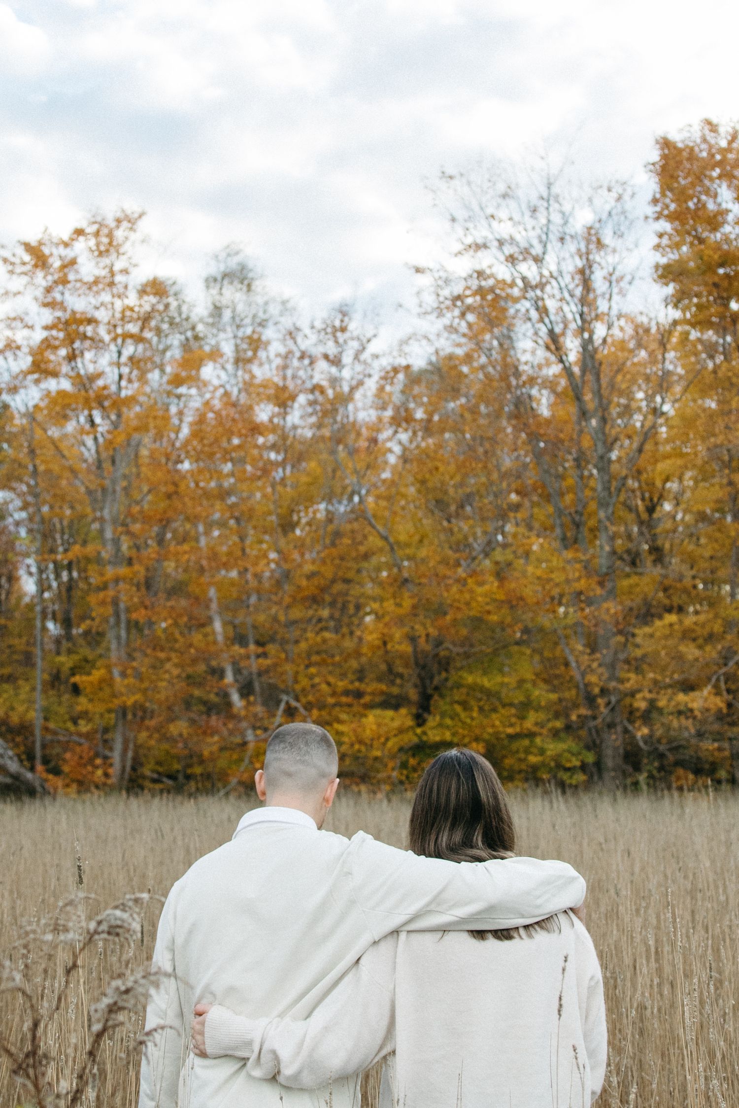 A couple in white sweaters embrace while looking at fall foliage in a tall grass field.