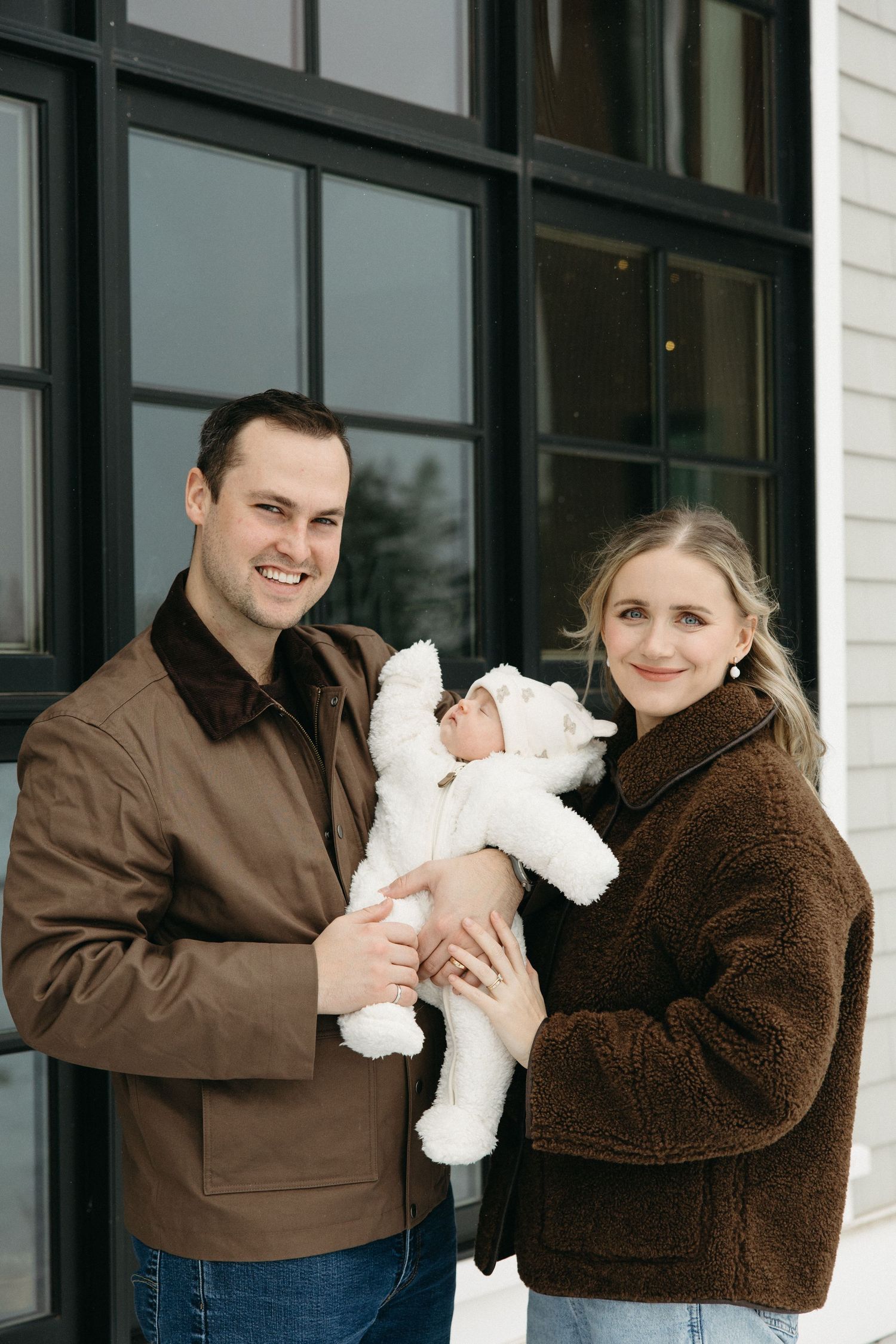 A couple poses with their newborn baby dressed in a fluffy white outfit outside a modern building.