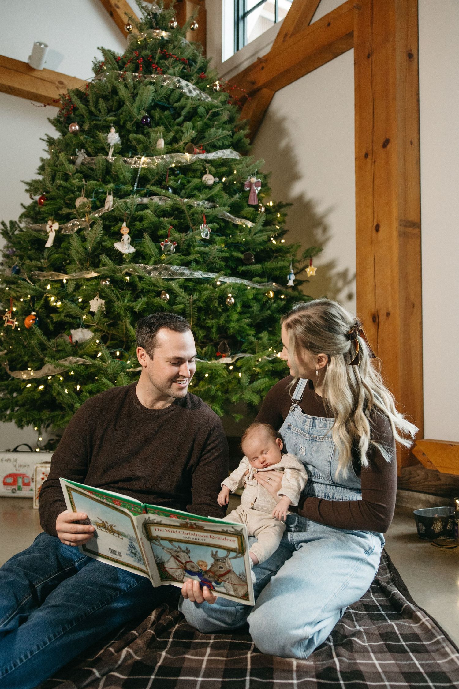 Family relaxing on plaid blanket while reading together by decorated Christmas tree.