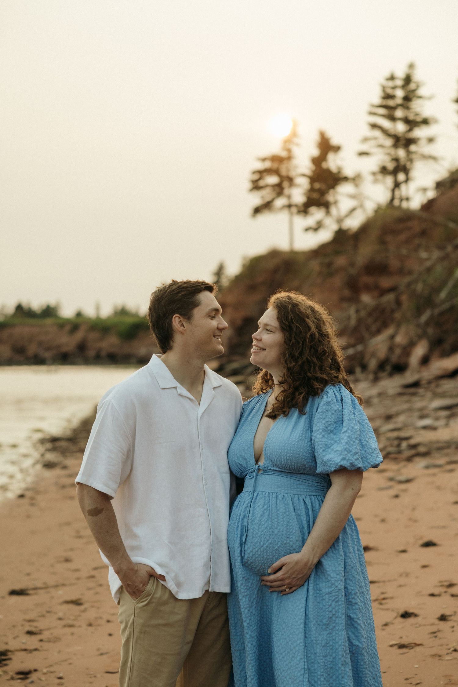 A couple in white and blue outfits share an intimate moment on a beach at sunset.