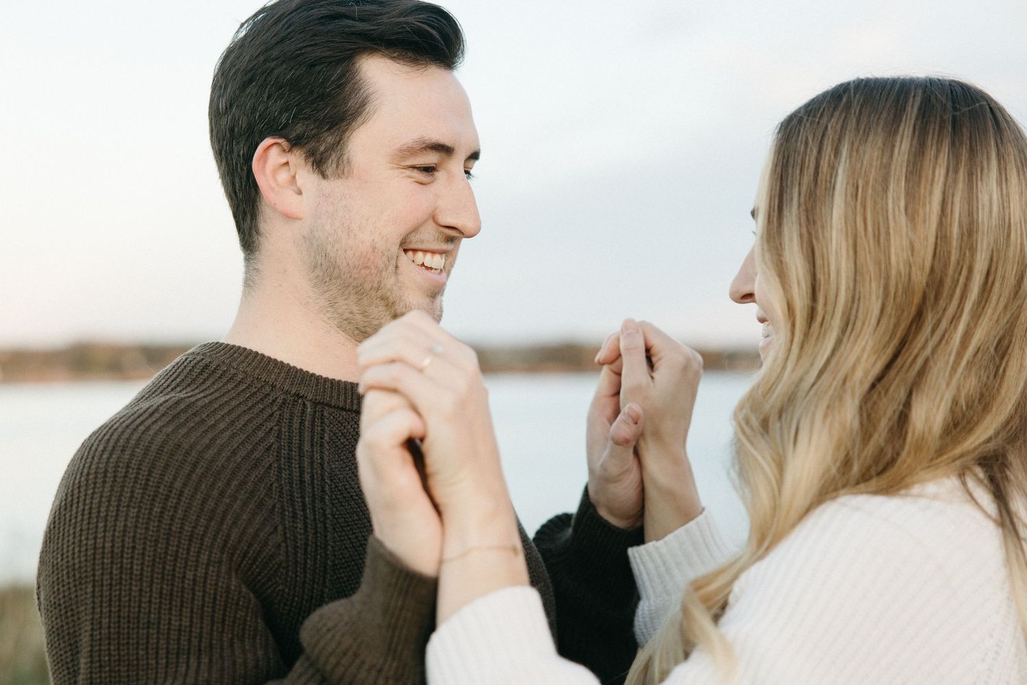 A smiling man in a brown sweater holds hands with a blonde woman in a white sweater during an outdoor couple portrait by the water.