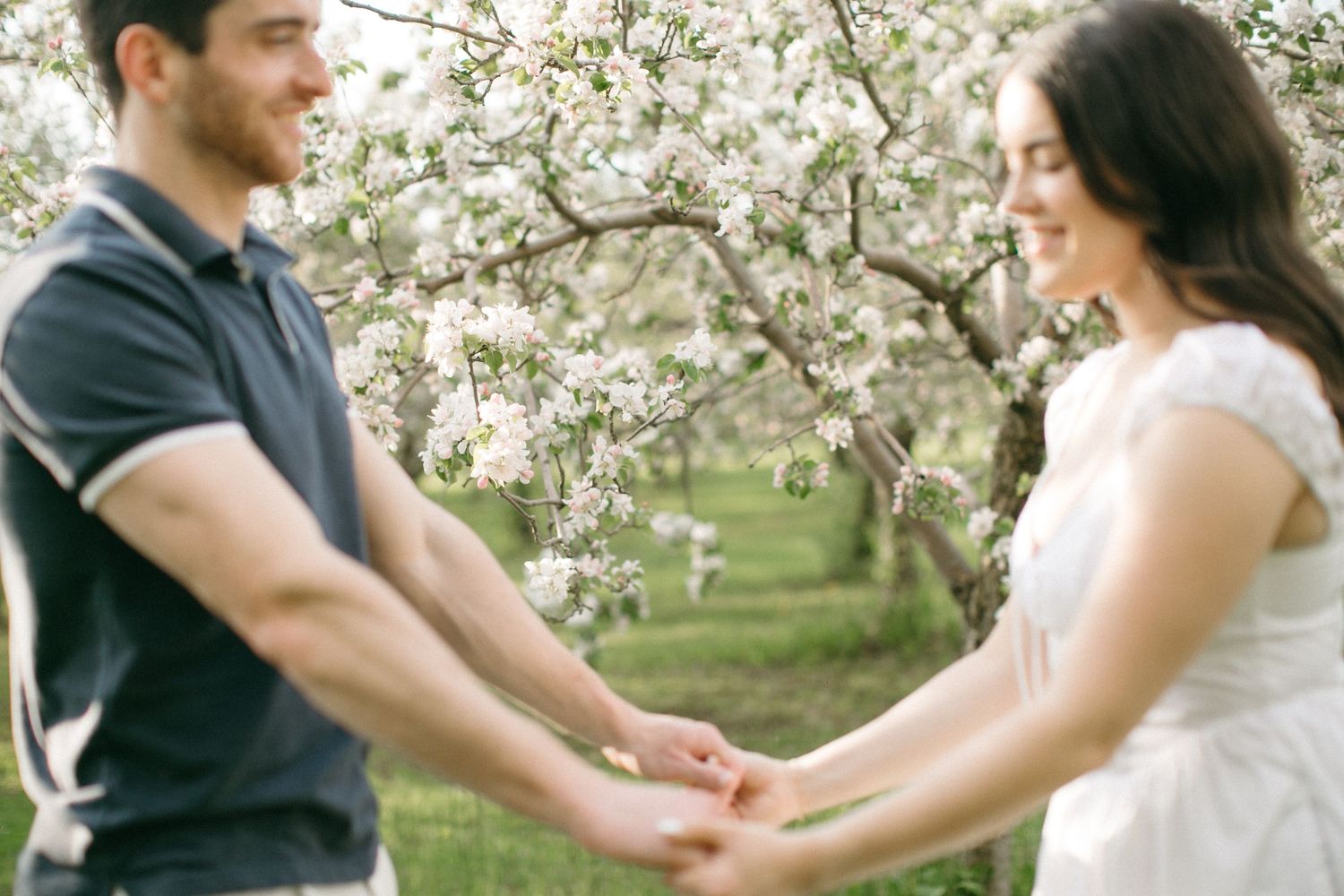 Couple holding hands and smiling in a blooming spring orchard with white flowering trees.