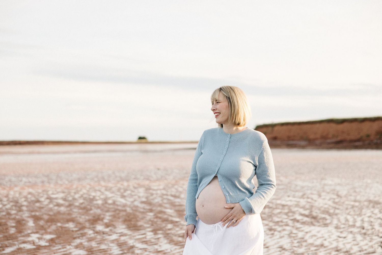 Smiling pregnant woman in blue sweater and white skirt holding her baby bump outdoors on a sandy landscape.