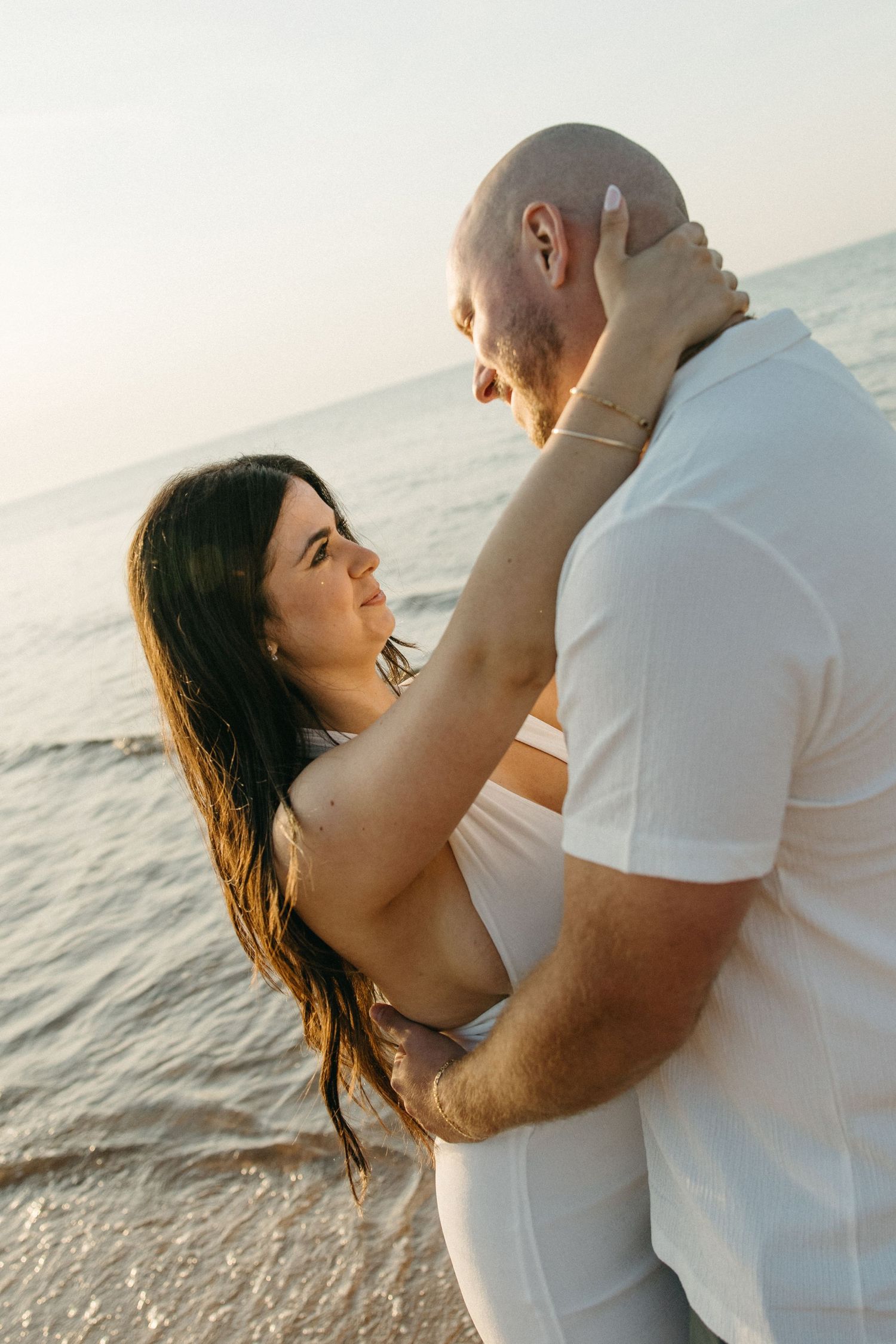 Romantic couple hugging by the ocean at sunset, woman in white dress gazing up at bald man.