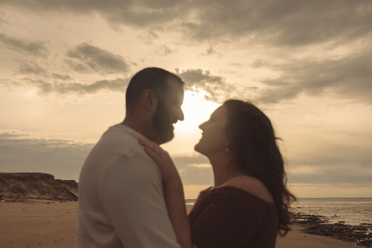 A couple walks along a scenic beach at sunset with dramatic cloudy skies overhead.