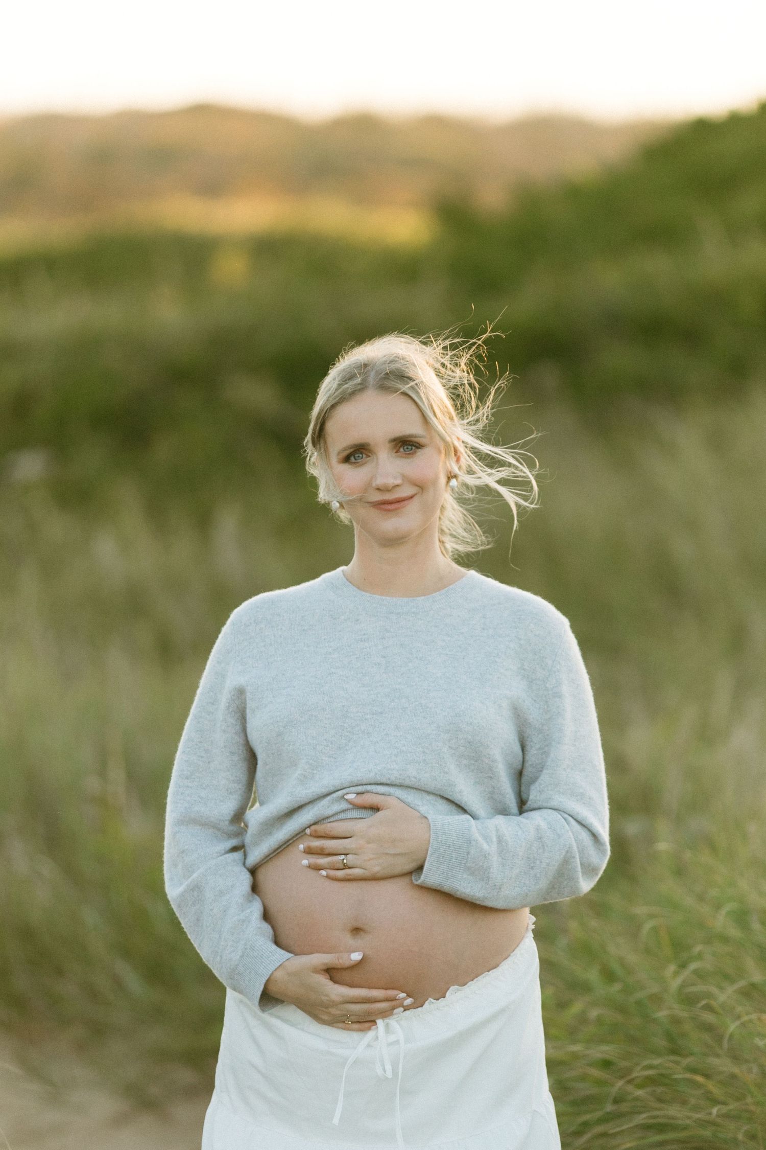 Expectant mother in gray sweater smiling while holding her belly in a grassy field at sunset.