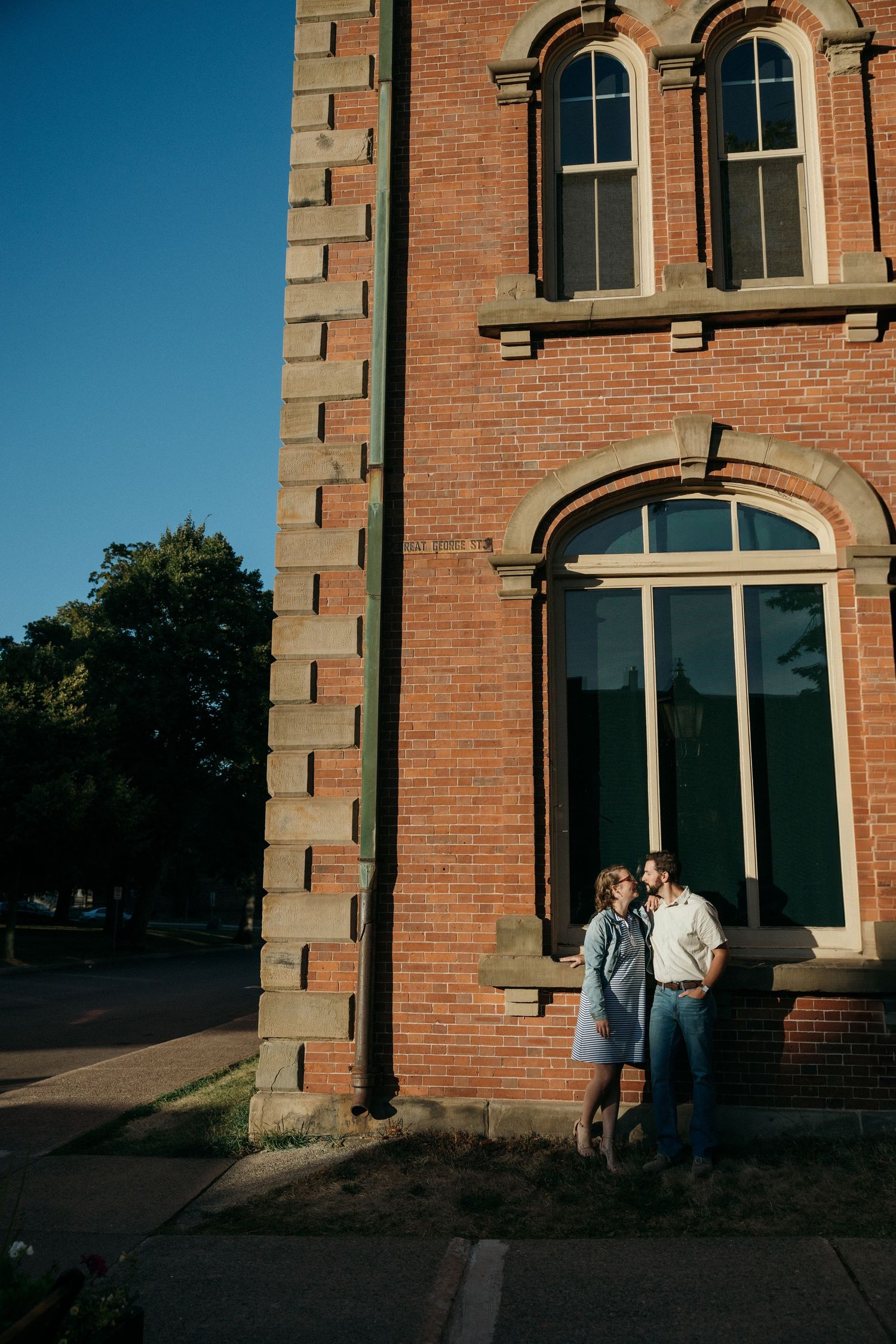 A couple poses against a red brick building with arched windows in golden evening light.