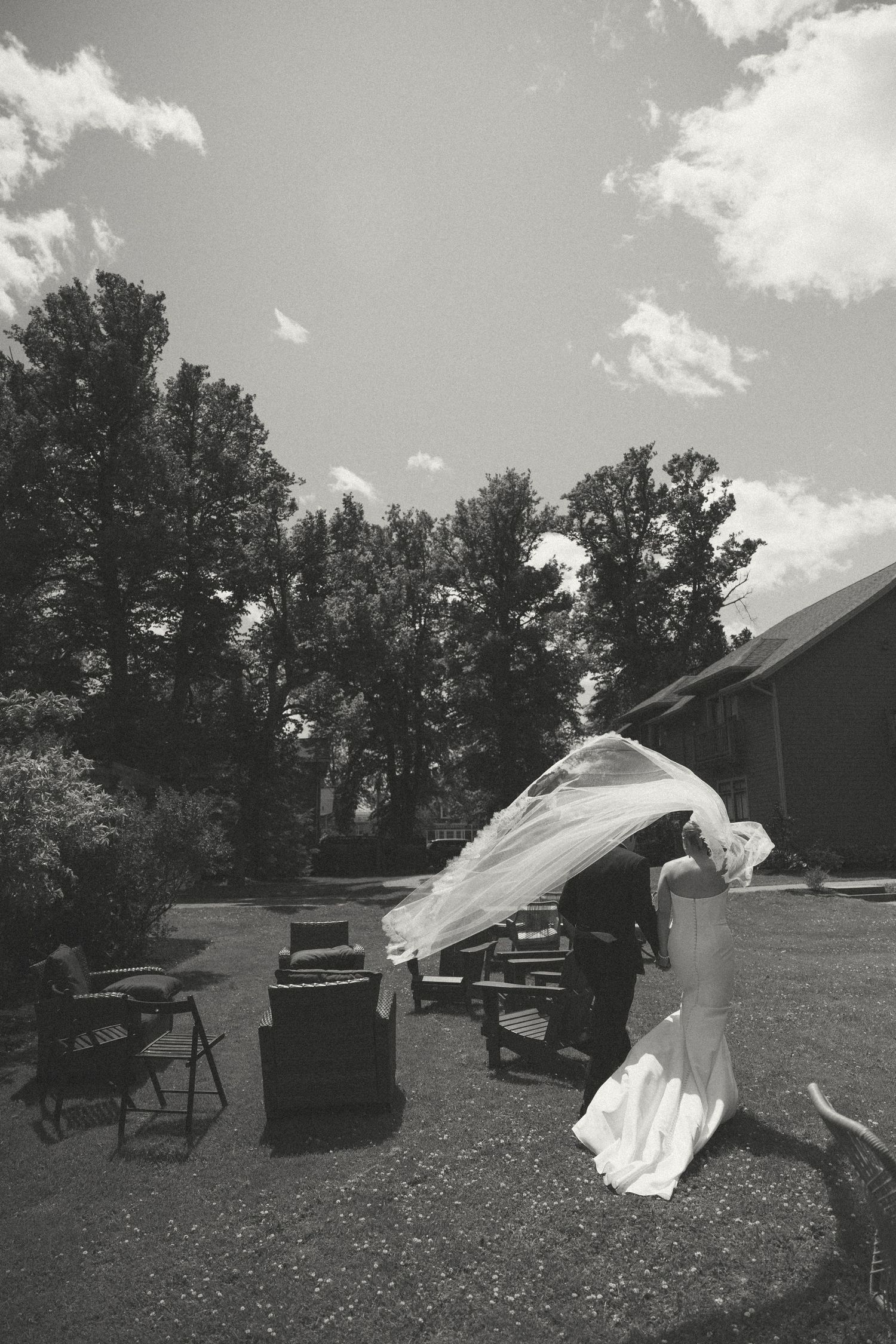 A flowing white wedding veil dramatically billows in the wind against a cloudy sky with trees in the background.