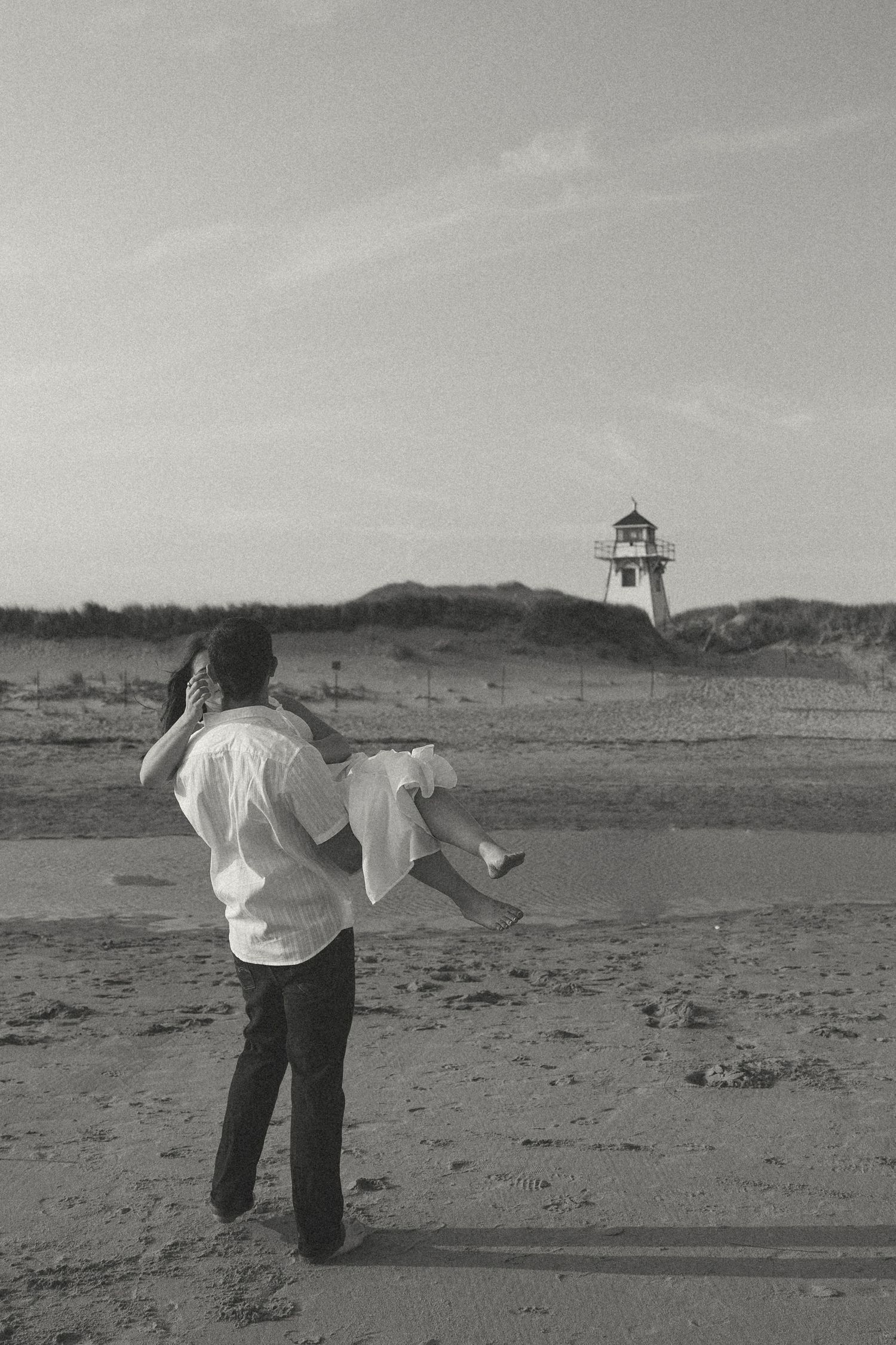 Black and white photo of people dancing on a beach with lighthouse in background.