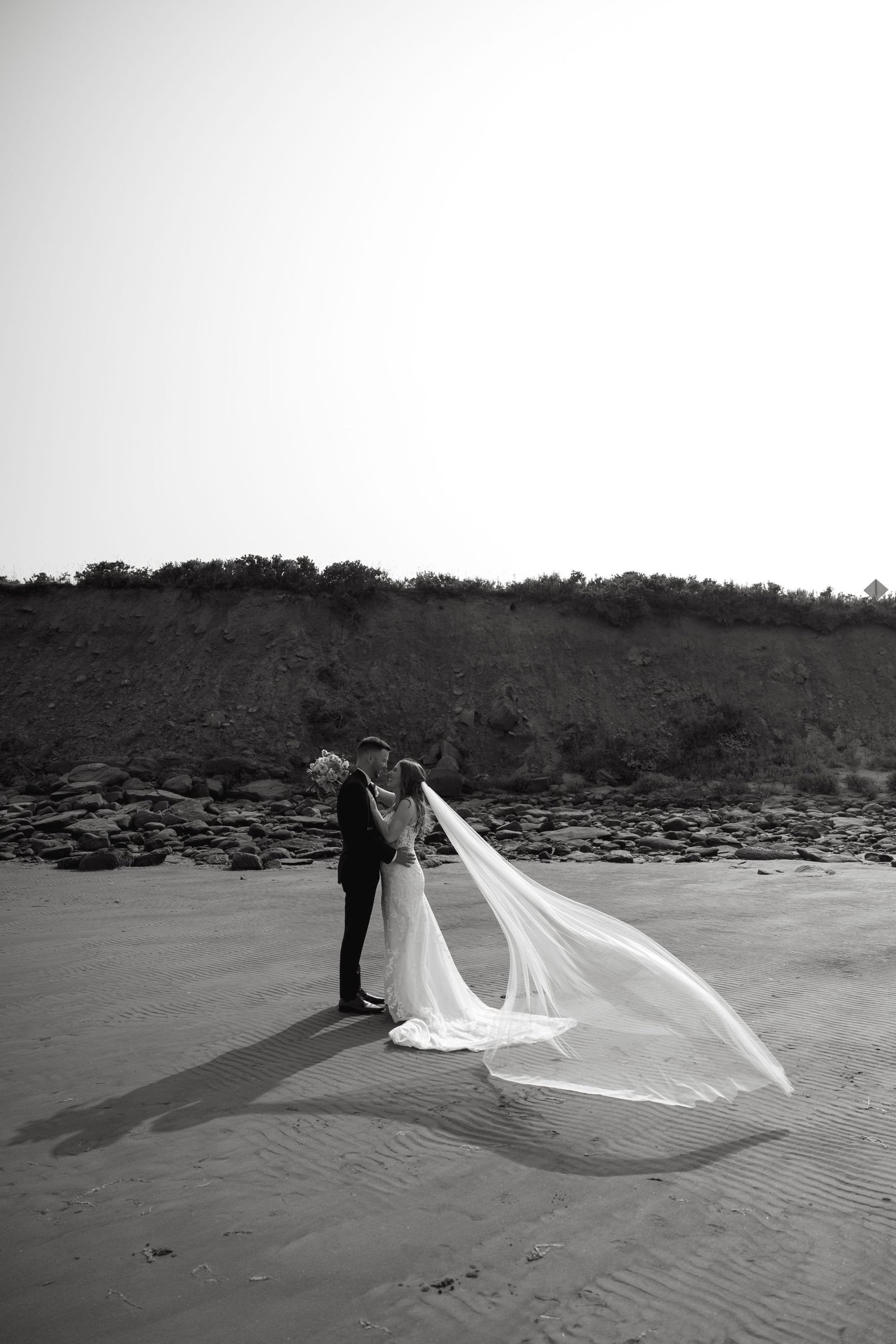 Black and white beach wedding portrait with dramatic veil movement on sandy shore.