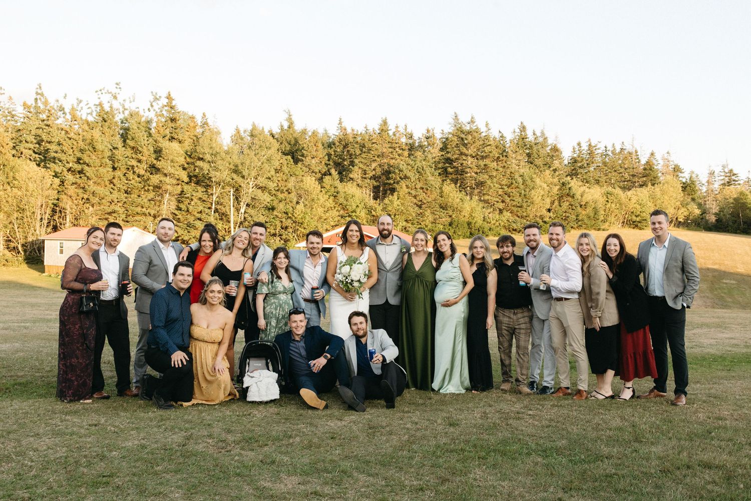 Large wedding party group photo with guests arranged on grassy hillside at sunset.