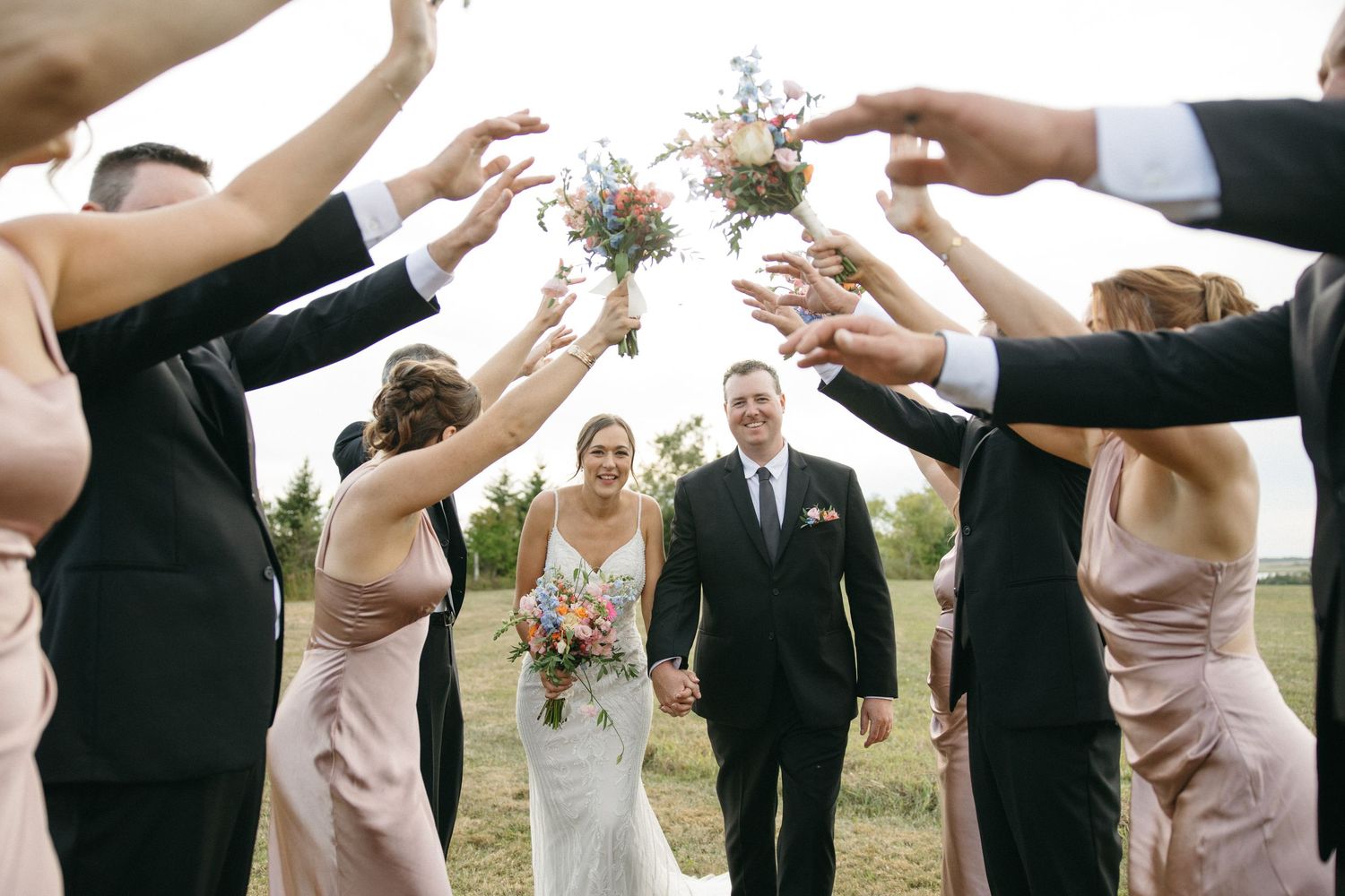 Wedding party forms an arch with their arms raised during an outdoor celebration.