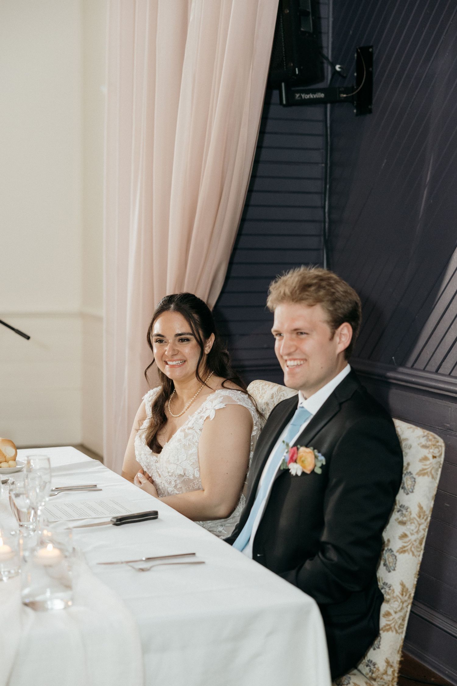 Couple sits at decorated wedding head table enjoying reception speeches.