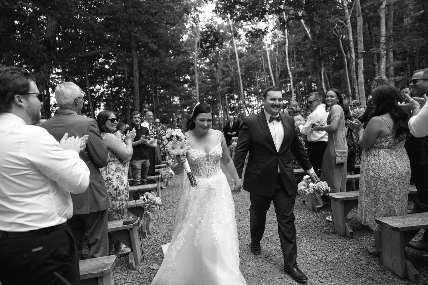 An outdoor wedding ceremony takes place in a wooded setting with guests watching.