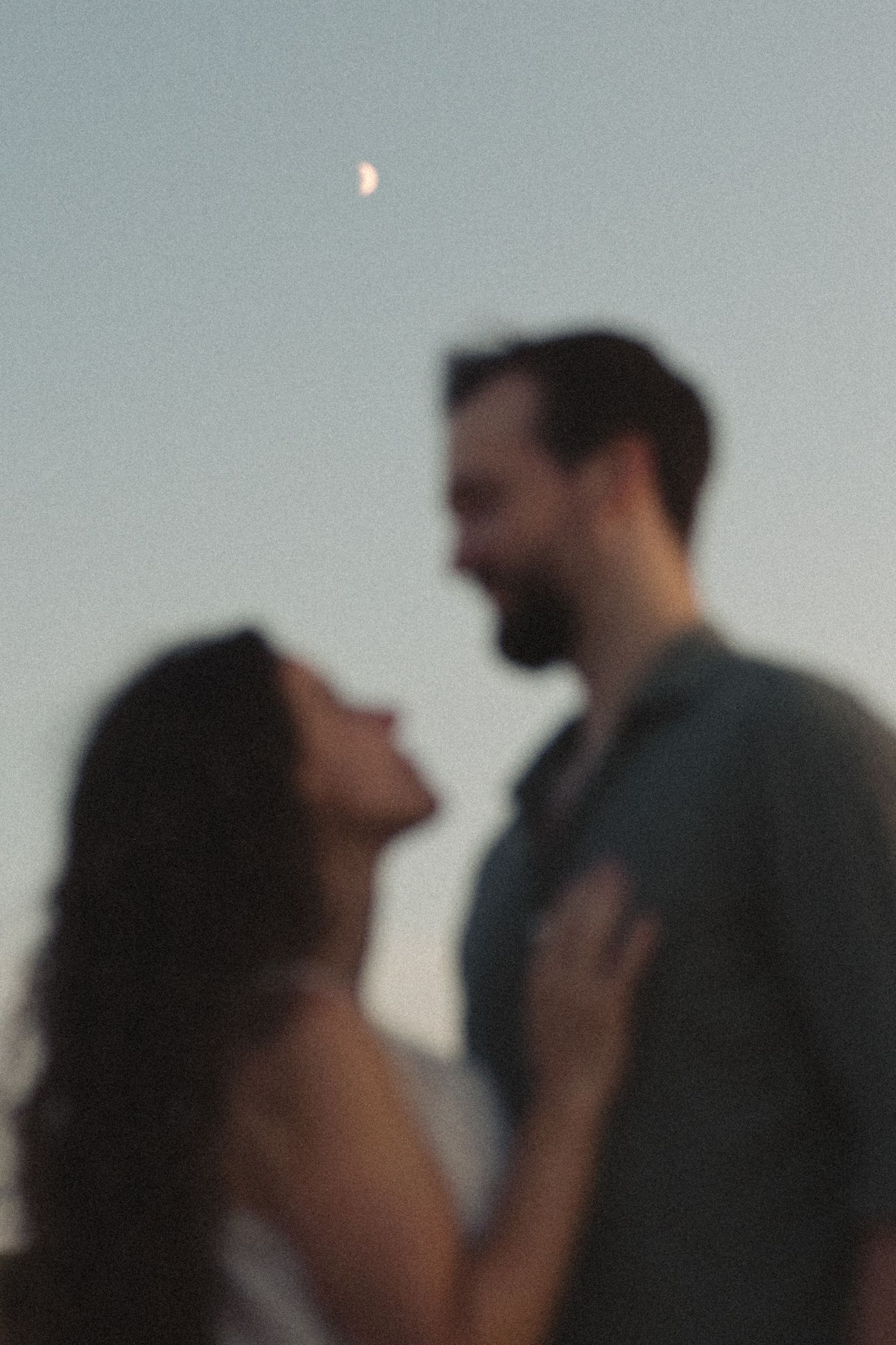 Soft focus romantic couple facing each other at dusk with crescent moon visible in twilight sky.