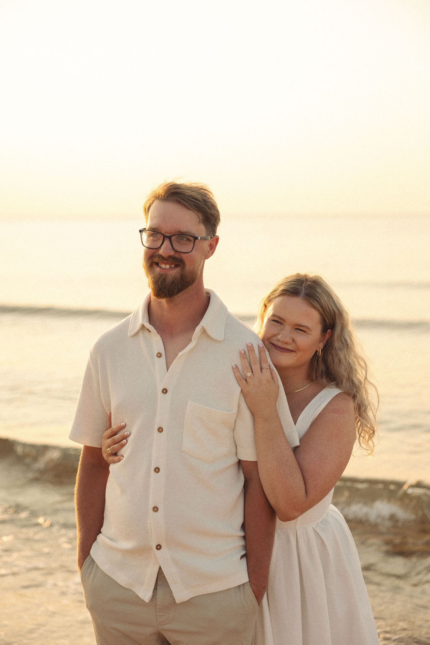 Smiling engaged couple posing at sunset on beach, woman showing off engagement ring in white dress.