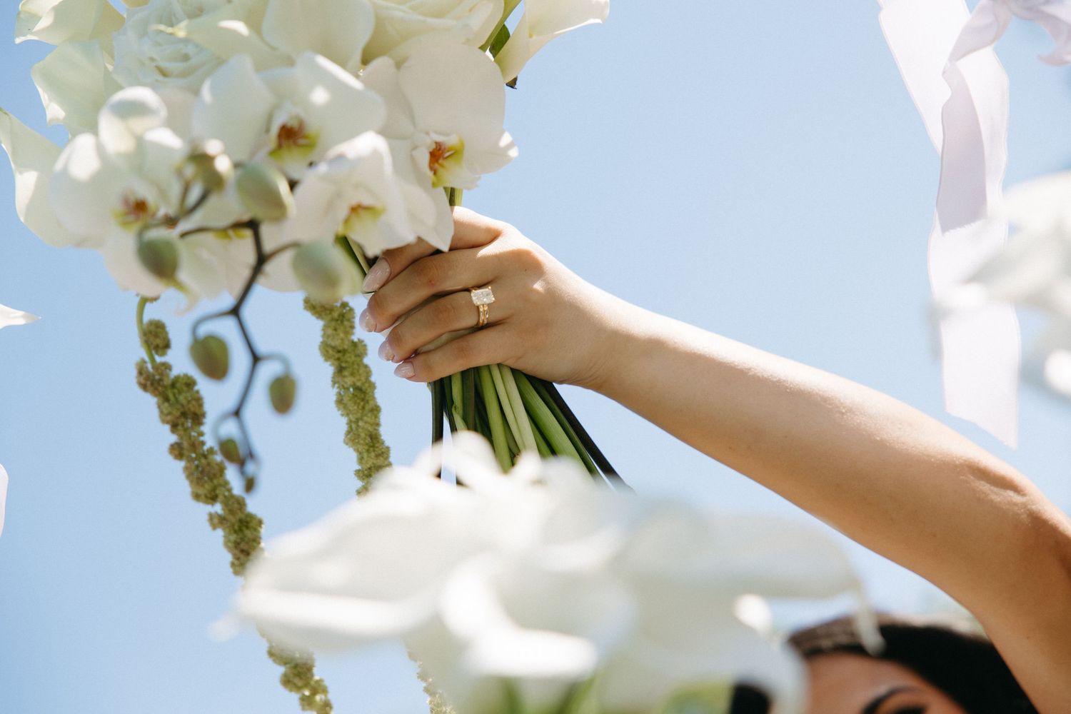 Bride holding a white orchid wedding bouquet against a clear blue sky, wearing a diamond engagement ring.