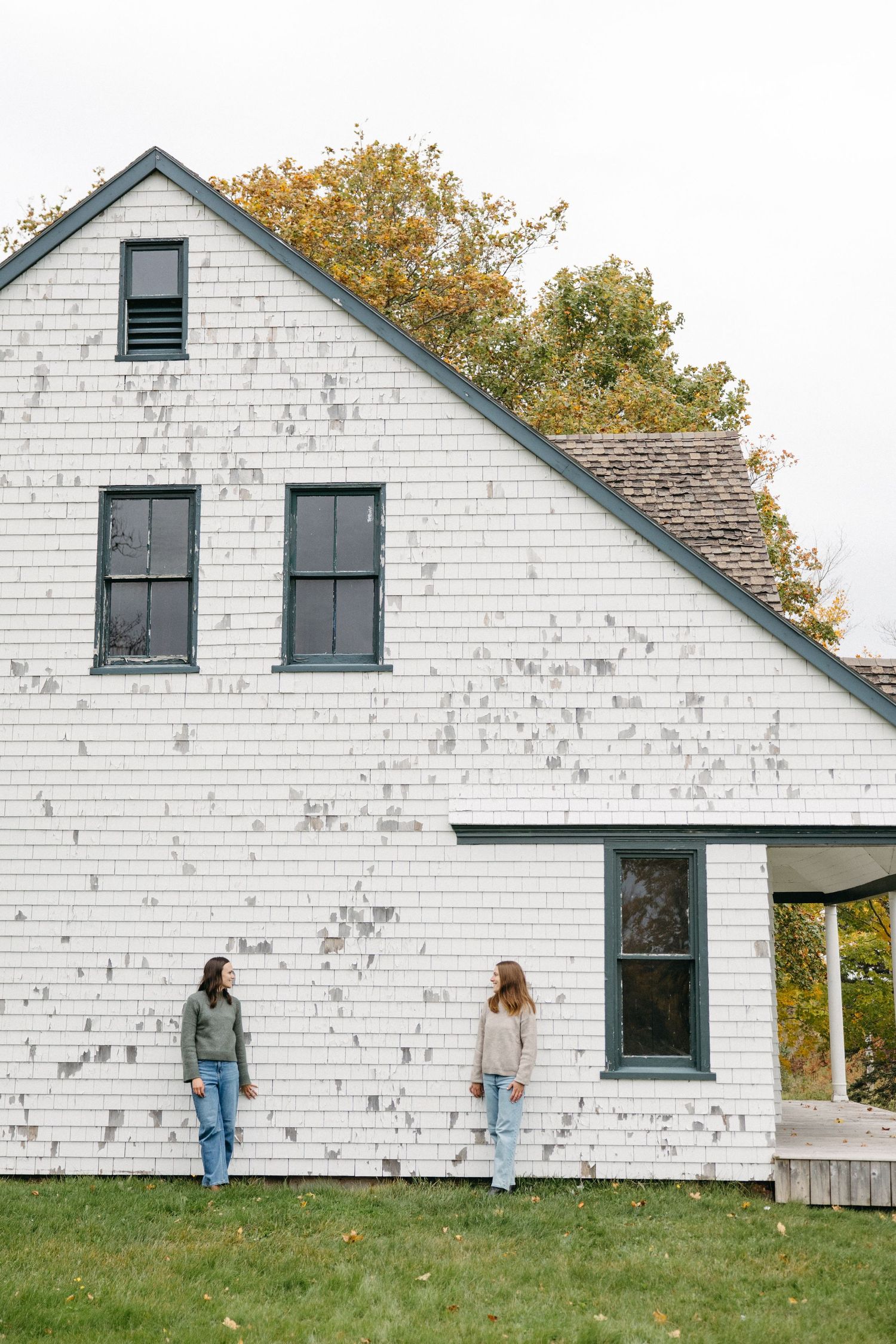 Two women in jeans and sweaters stand on a green lawn beside a weathered white shingled farmhouse with dark windows and autumn trees behind.