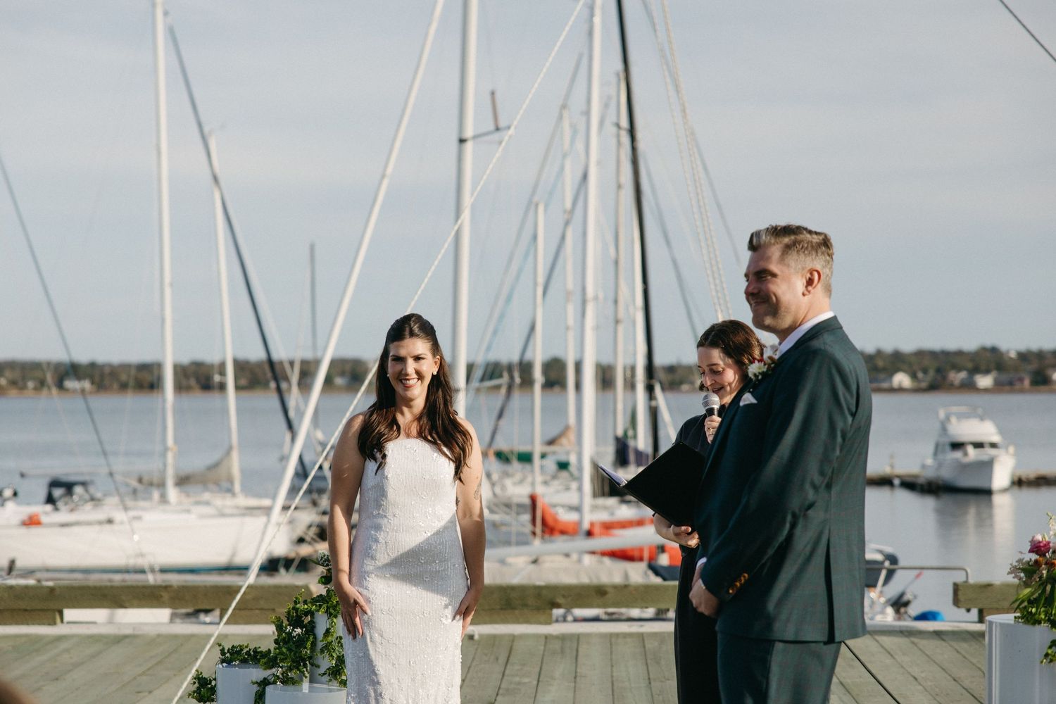 Smiling bride in a white dress and groom in a dark suit stand with an officiant at a waterfront wedding ceremony beside sailboats.