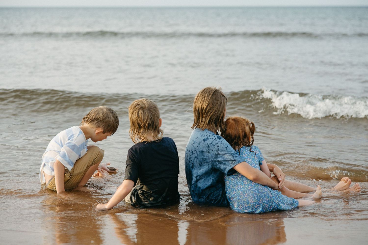 Four children sitting together at the water's edge on a sunny beach.