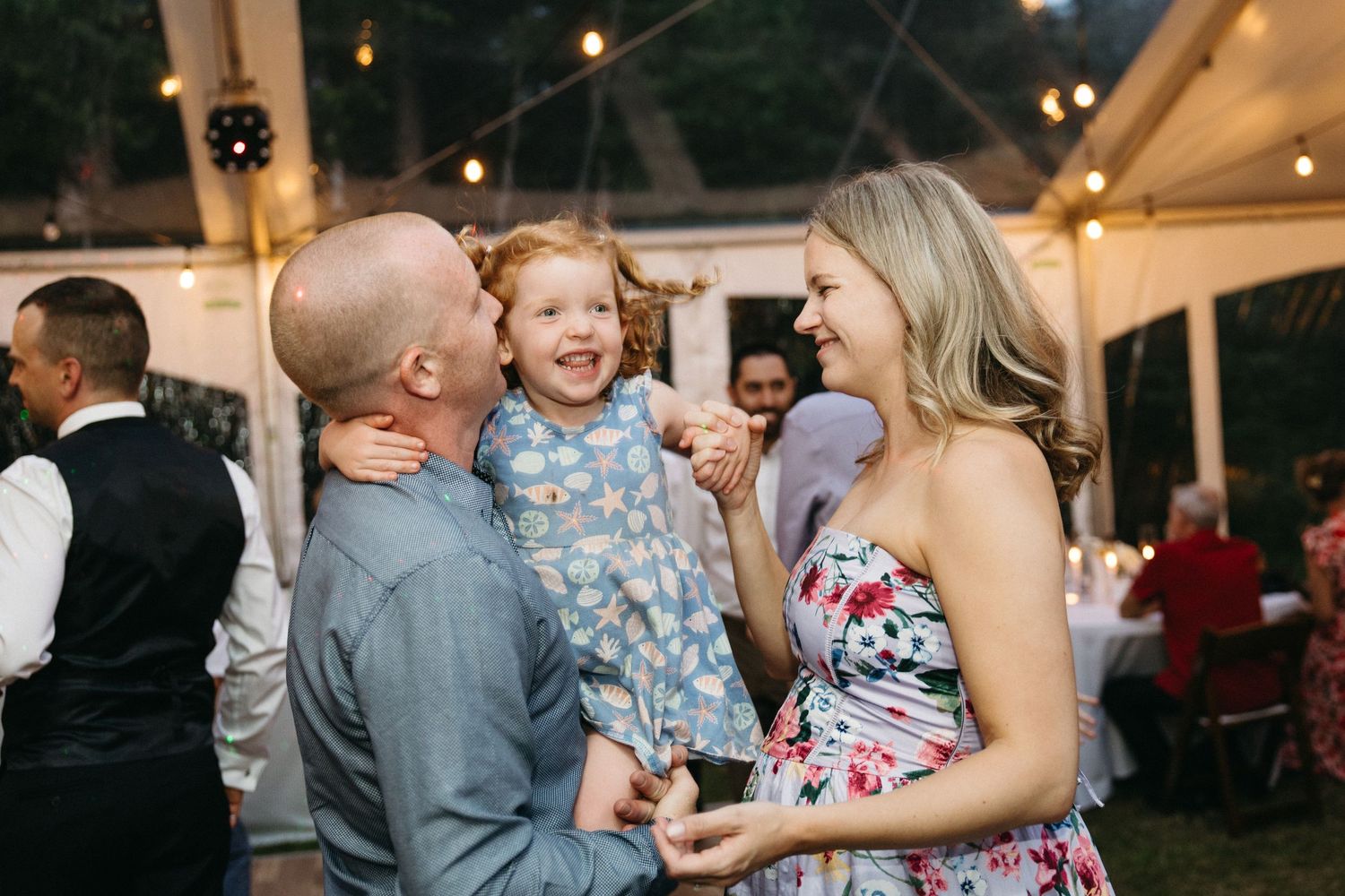 Joyful family moment as parents kiss and laugh with their little girl at a wedding reception.