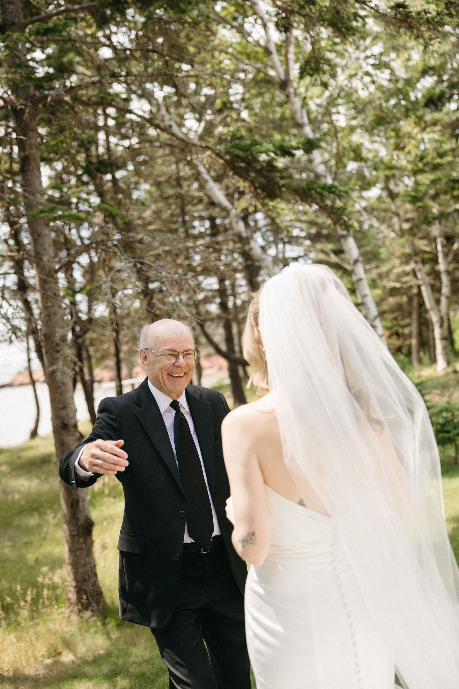 Emotional father smiles reaching toward daughter bride in white gown during first look outdoors.