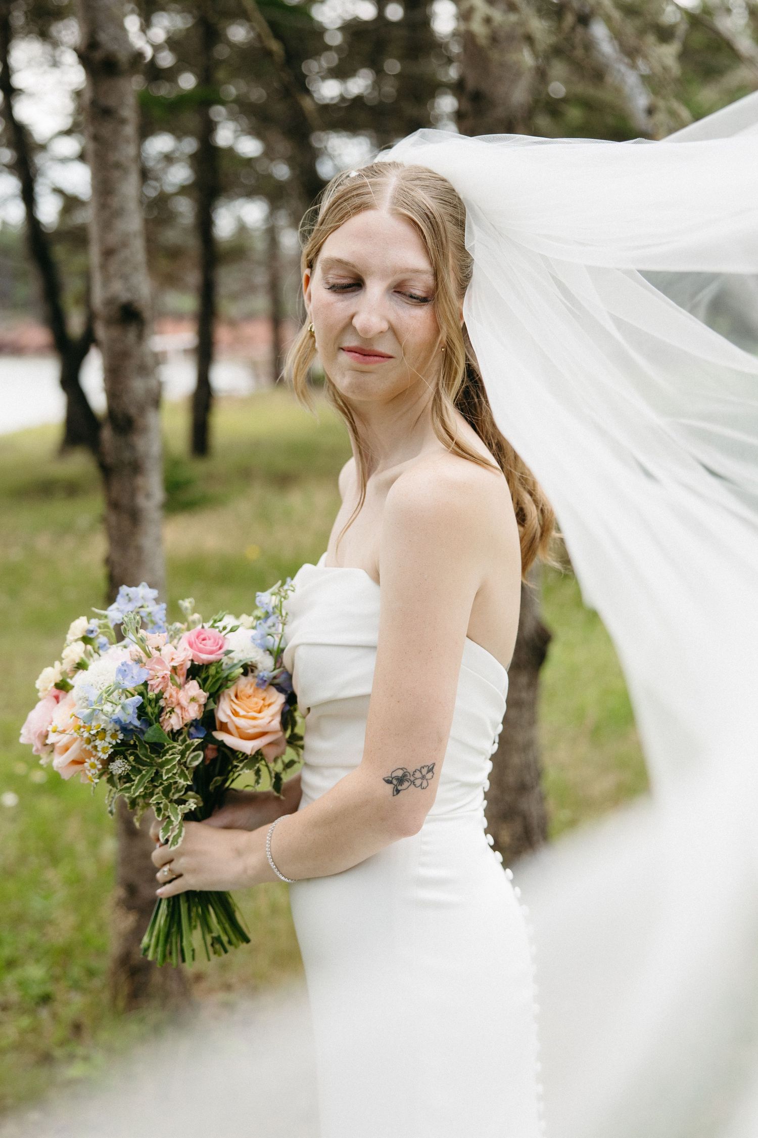 Bride in strapless white gown holds colorful bouquet as veil flows in the wind outdoors.