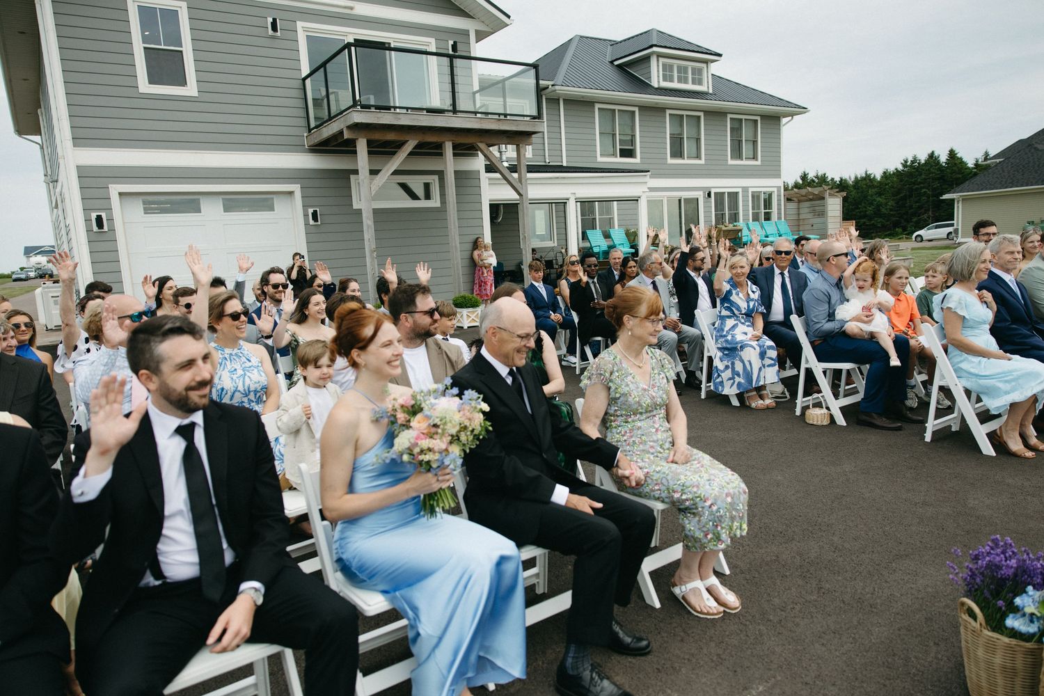 Large outdoor wedding ceremony with guests seated in white chairs in front of coastal gray home.