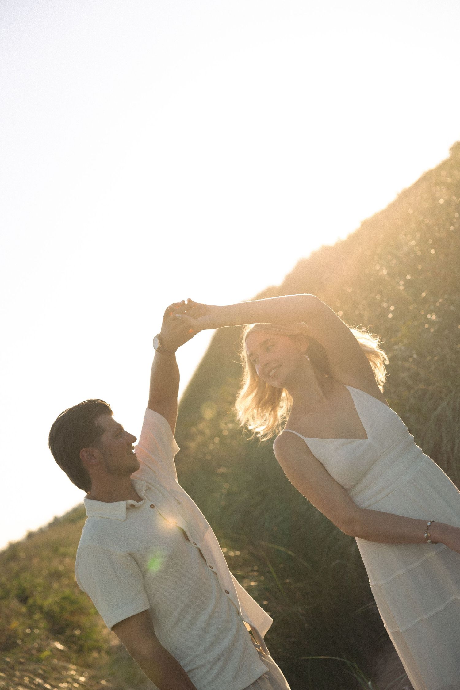 Couple dancing outdoors in golden sunset light, man spinning woman in white dress on hillside.
