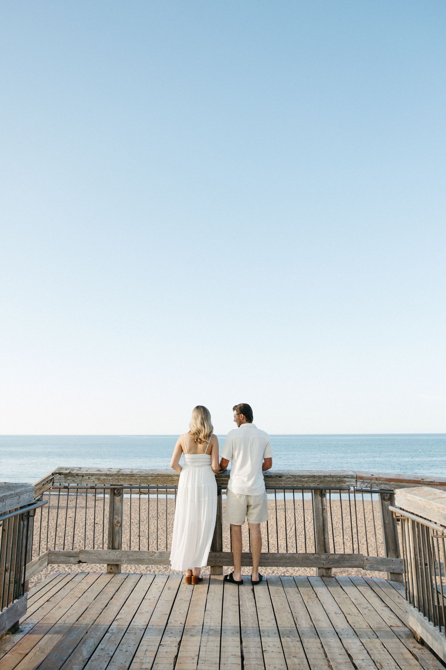 Couple in white outfits standing on wooden boardwalk overlooking calm ocean under clear blue sky.