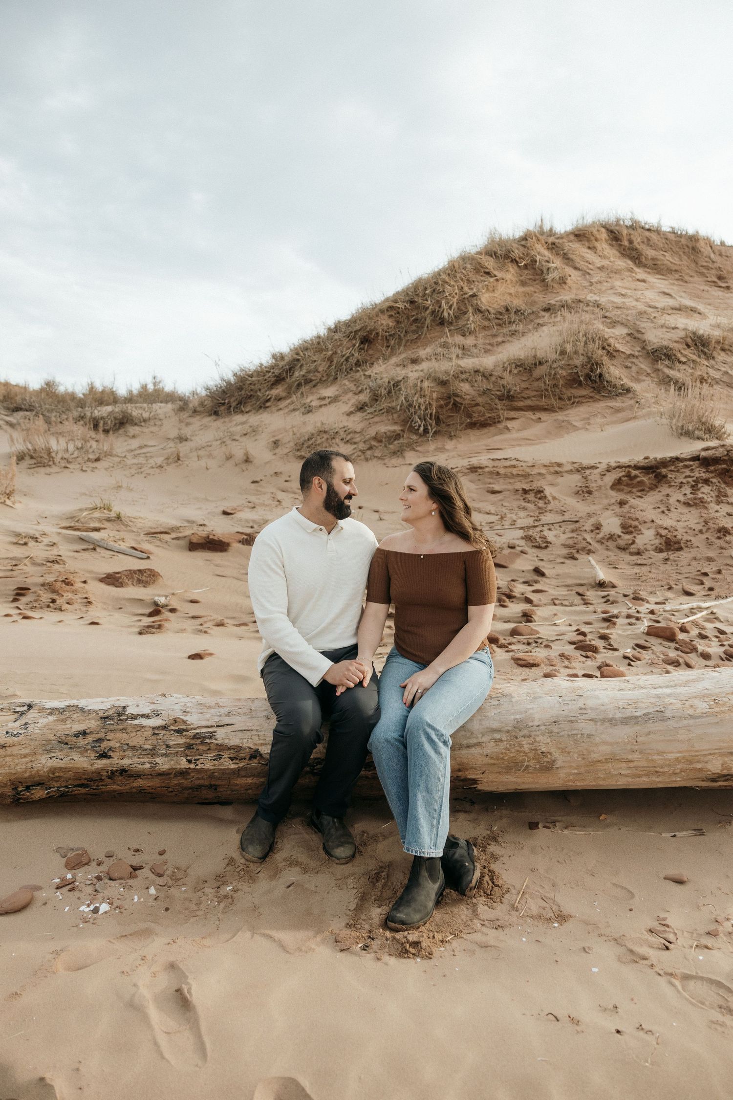 Two people sitting together on a weathered log among coastal sand dunes.