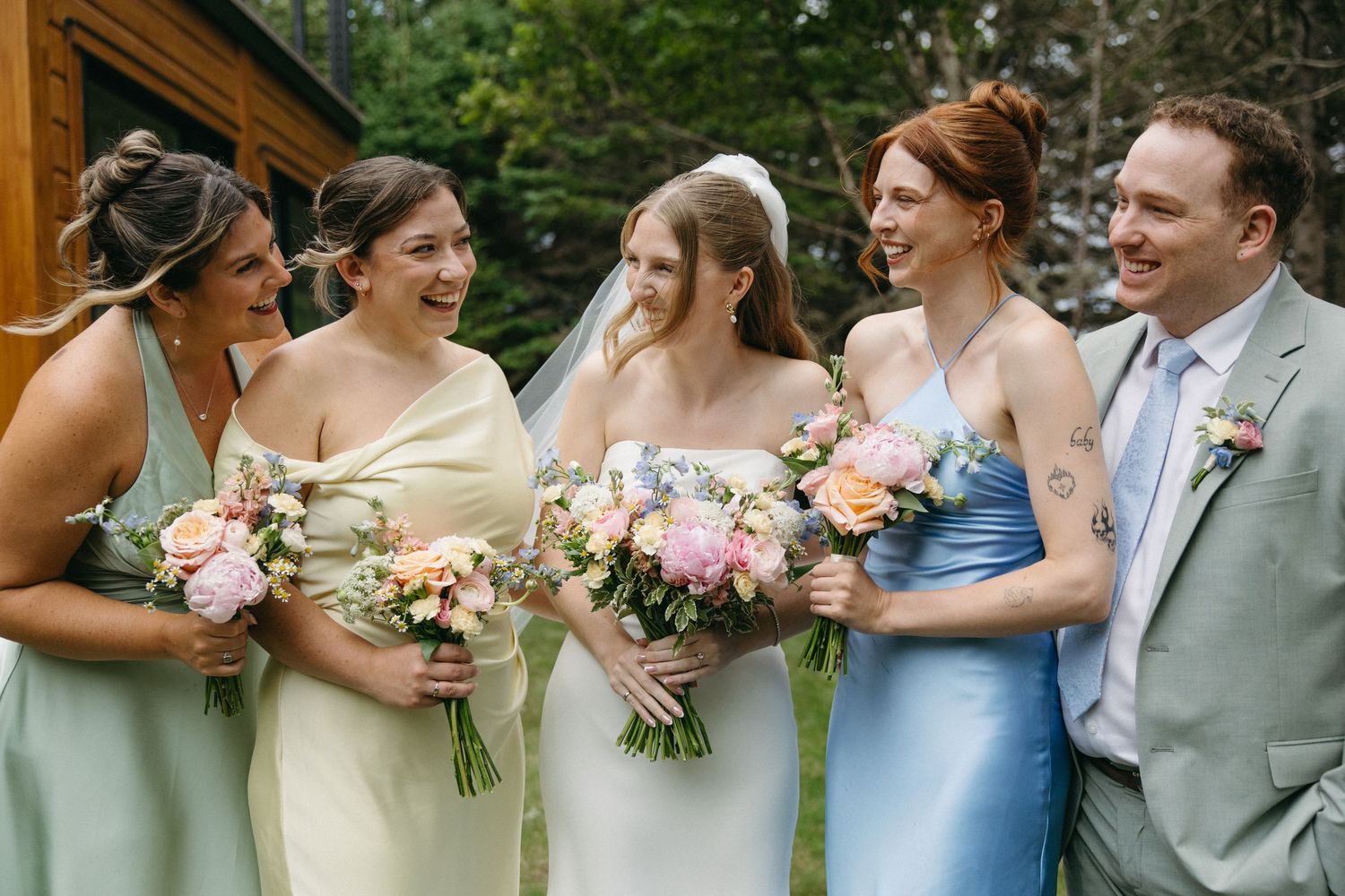 Bride in white gown laughs with bridesmaids in pastel dresses and groomsman in gray suit outdoors.