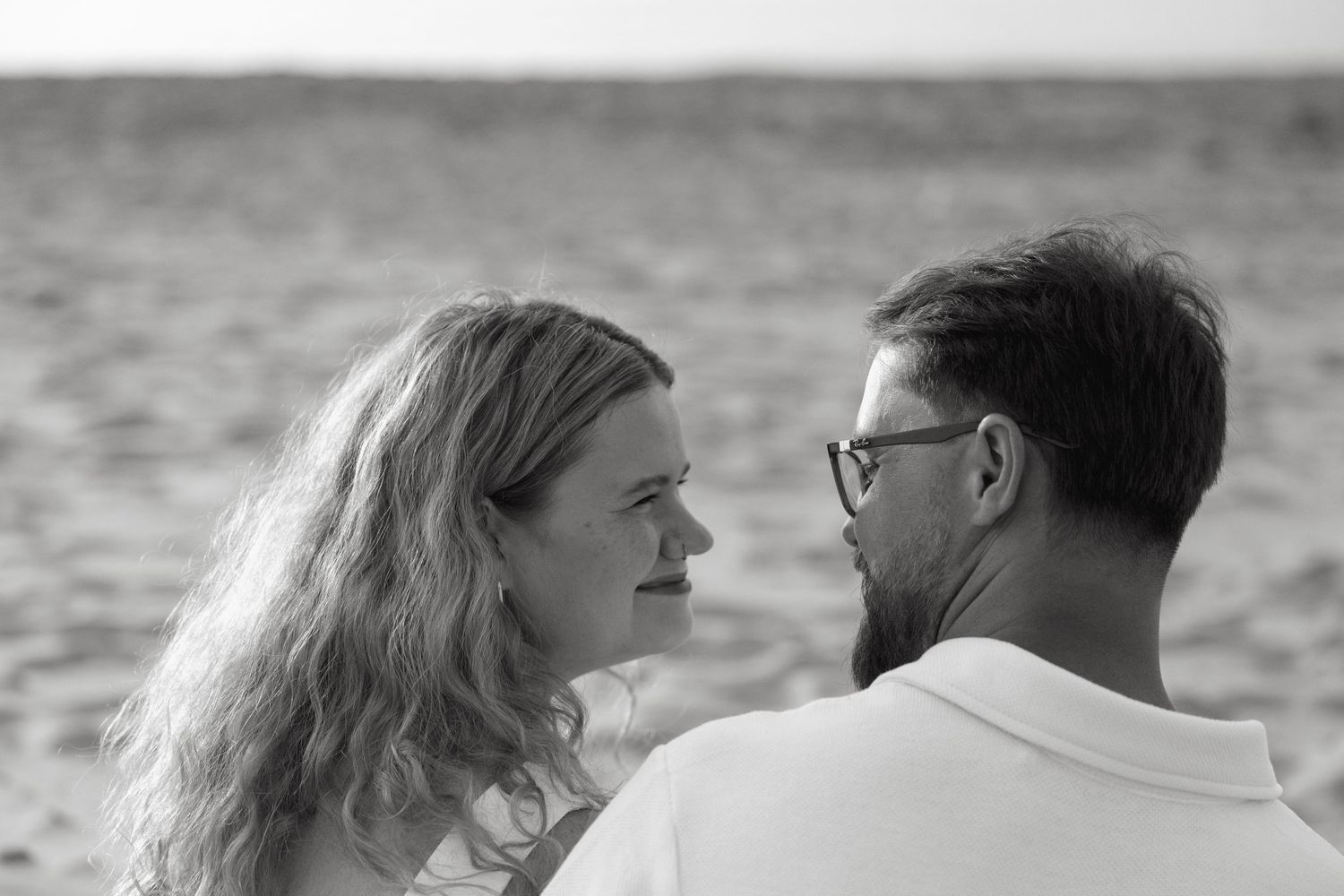 Black and white photo of a couple gazing at each other on a beach, with ocean waves in the background.