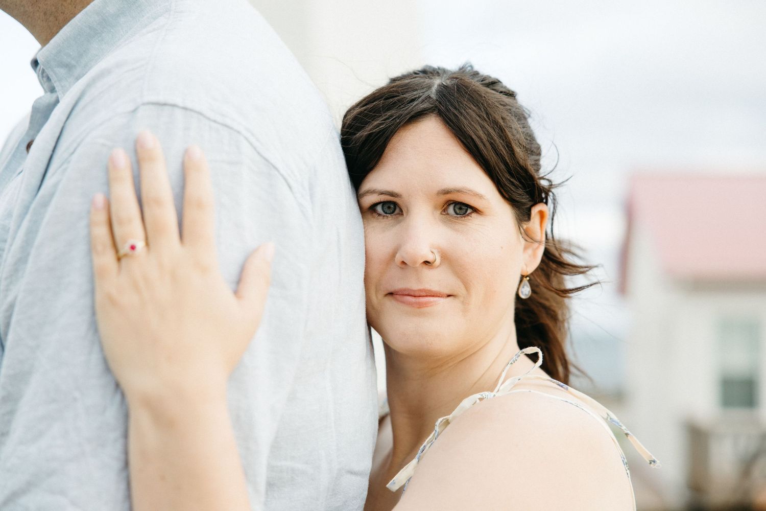 Bride in white veil leans against groom in gray suit, showcasing wedding ring, with soft outdoor background.