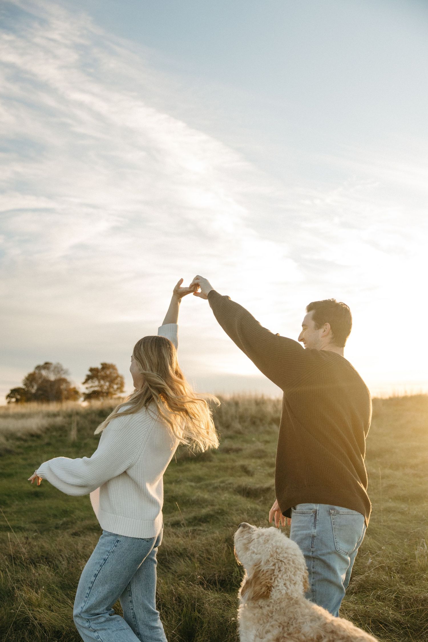 Couple dancing hand in hand in a golden field at sunset while a fluffy dog watches, capturing a joyful outdoor engagement moment.