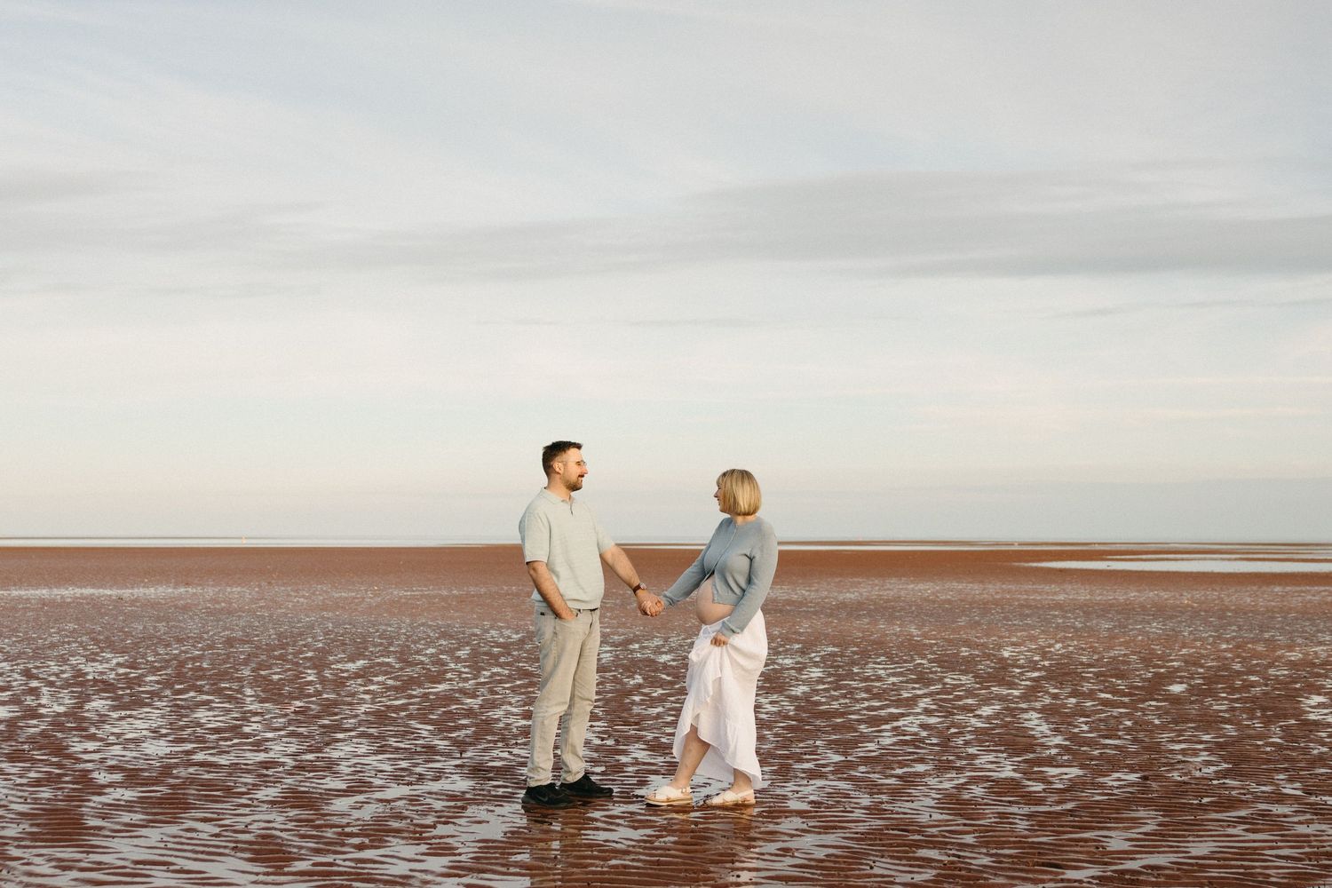 Couple holding hands facing each other on a vast red sandy beach under a cloudy sky.