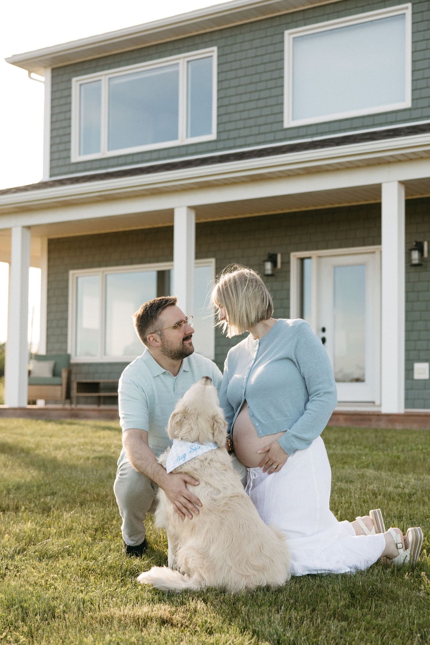 Happy pregnant couple with golden retriever dog sitting on grass in front of modern green home at sunset.