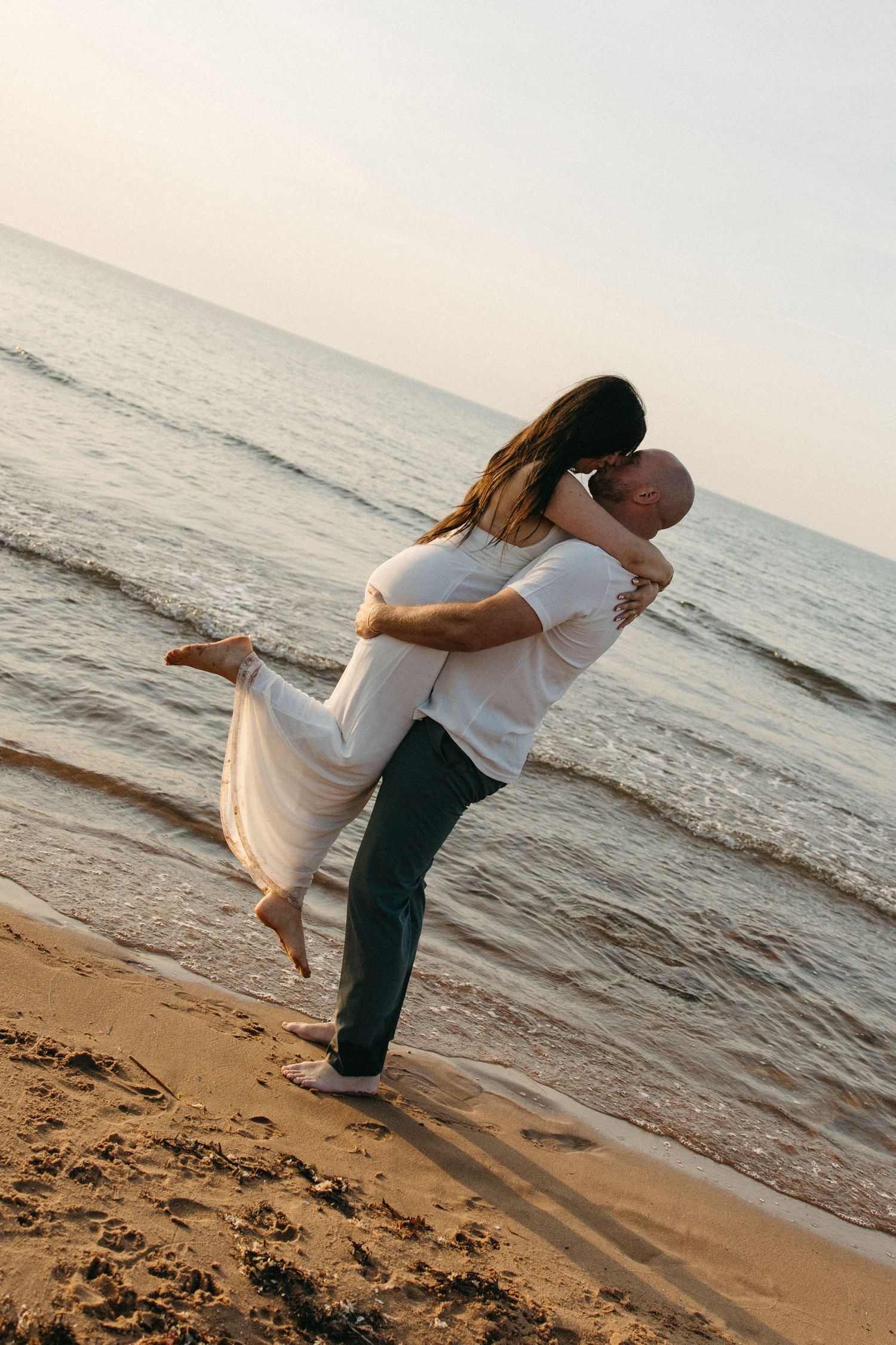 Couple embracing on sandy beach at sunset, man lifting woman in white dress near ocean waves.