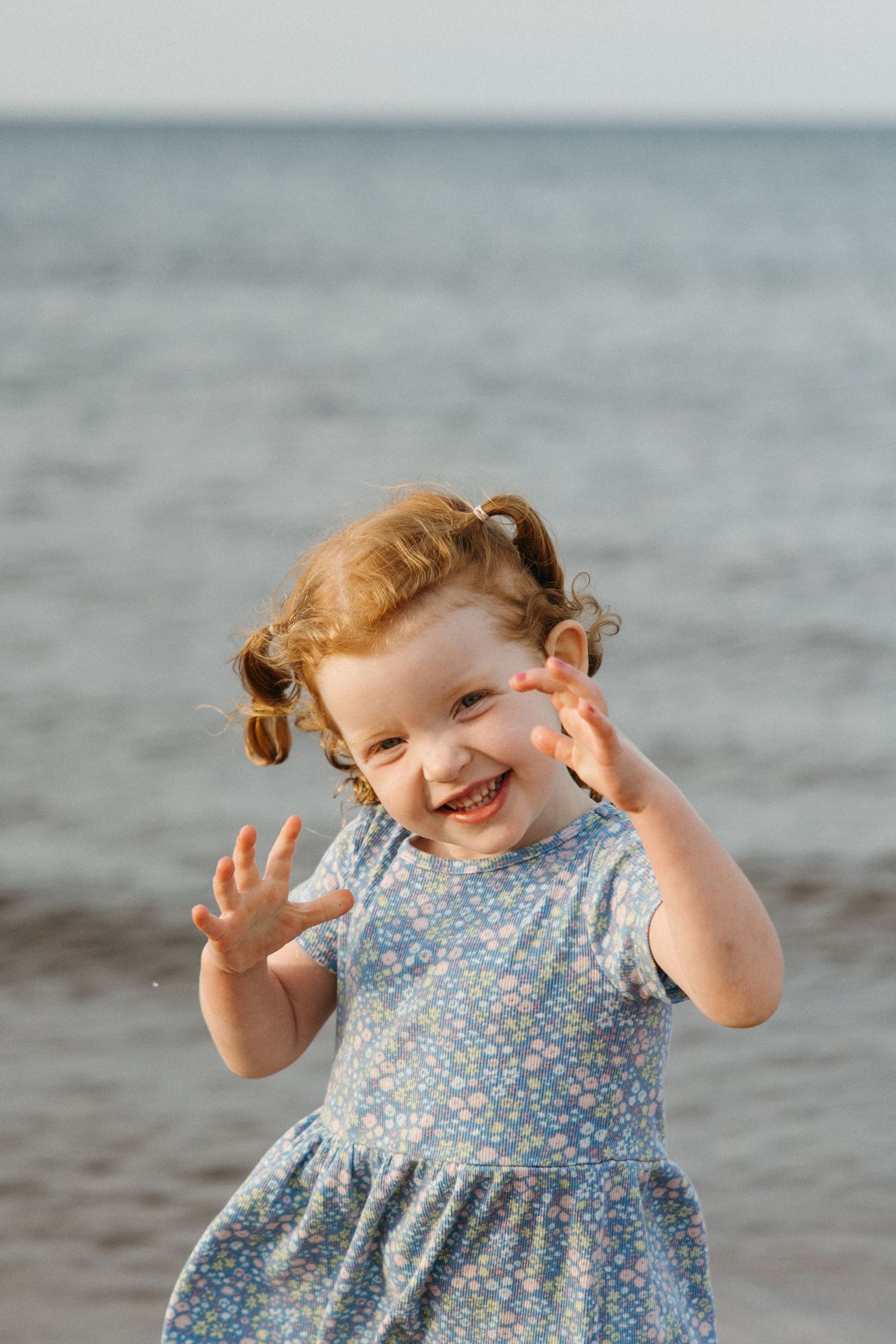 A joyful toddler girl with pigtails in a floral blue dress laughs and raises her hands at the beach.