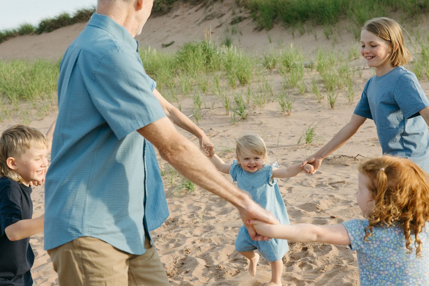 Family holding hands in a circle with young toddler on sunny sandy beach.