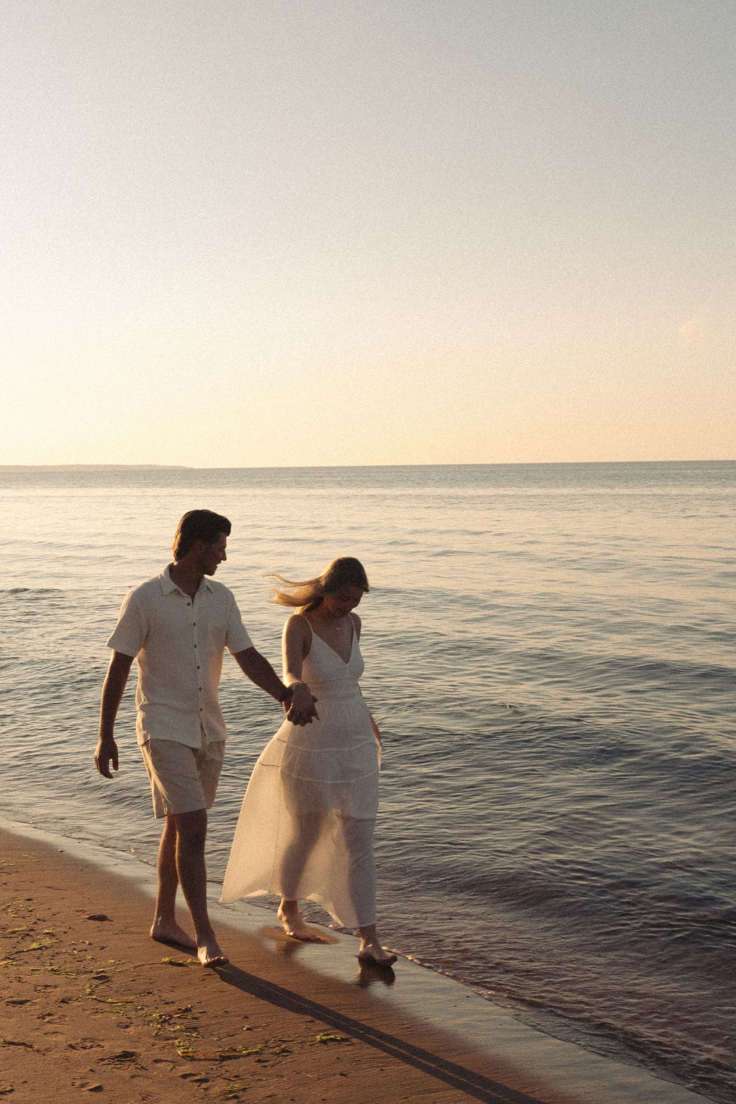 Couple holding hands walking along sandy beach shore at sunset.
