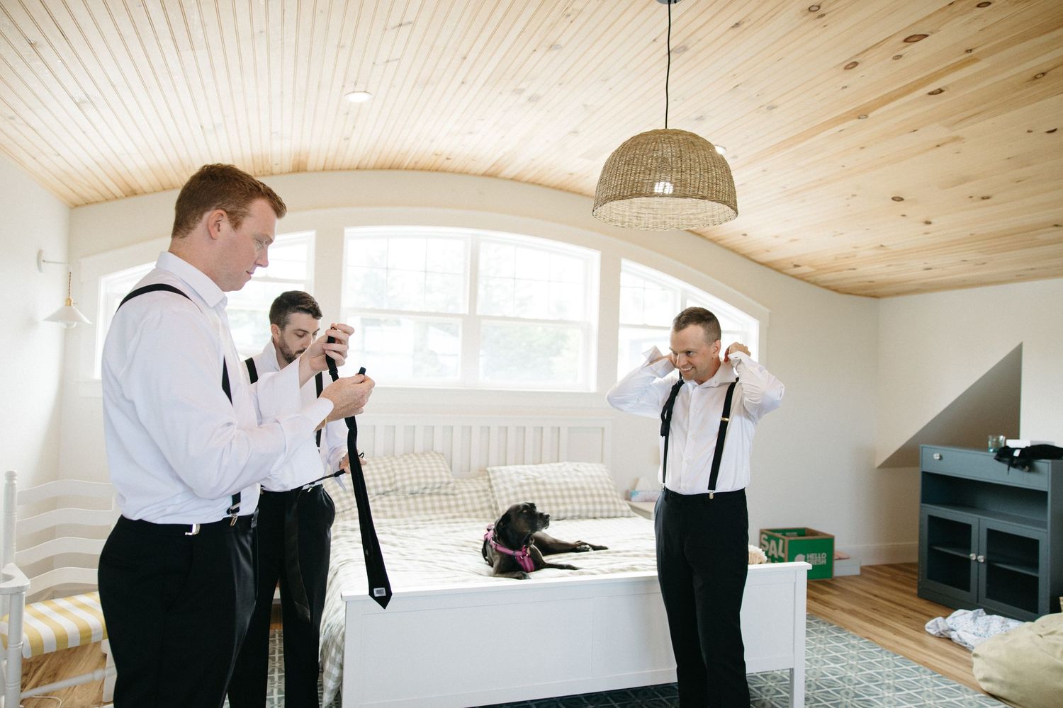 Groomsmen in white shirts and suspenders adjusting ties in rustic wood-paneled bedroom with dog on bed.