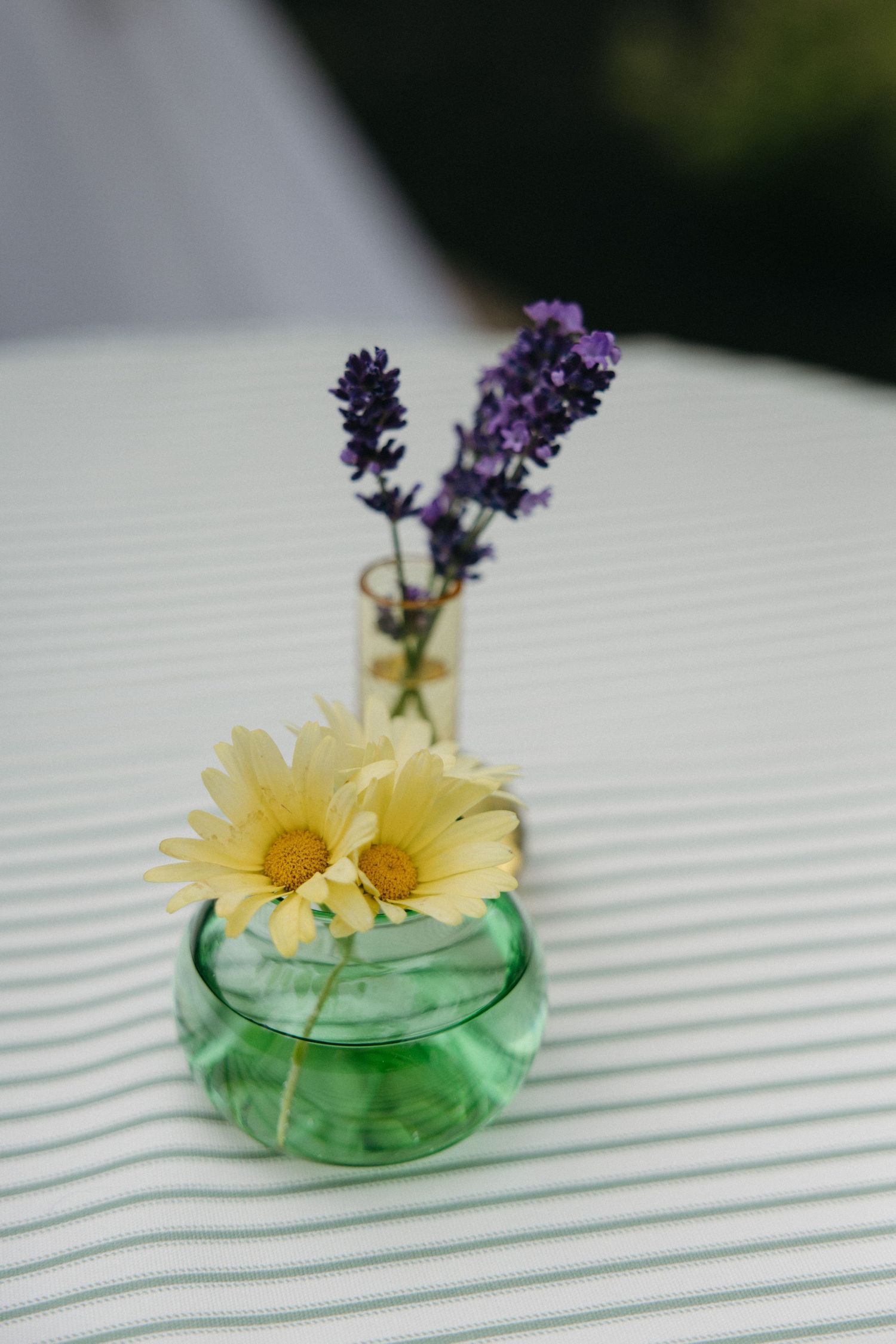 Yellow daisies and purple lavender in small green glass vase on striped tablecloth.