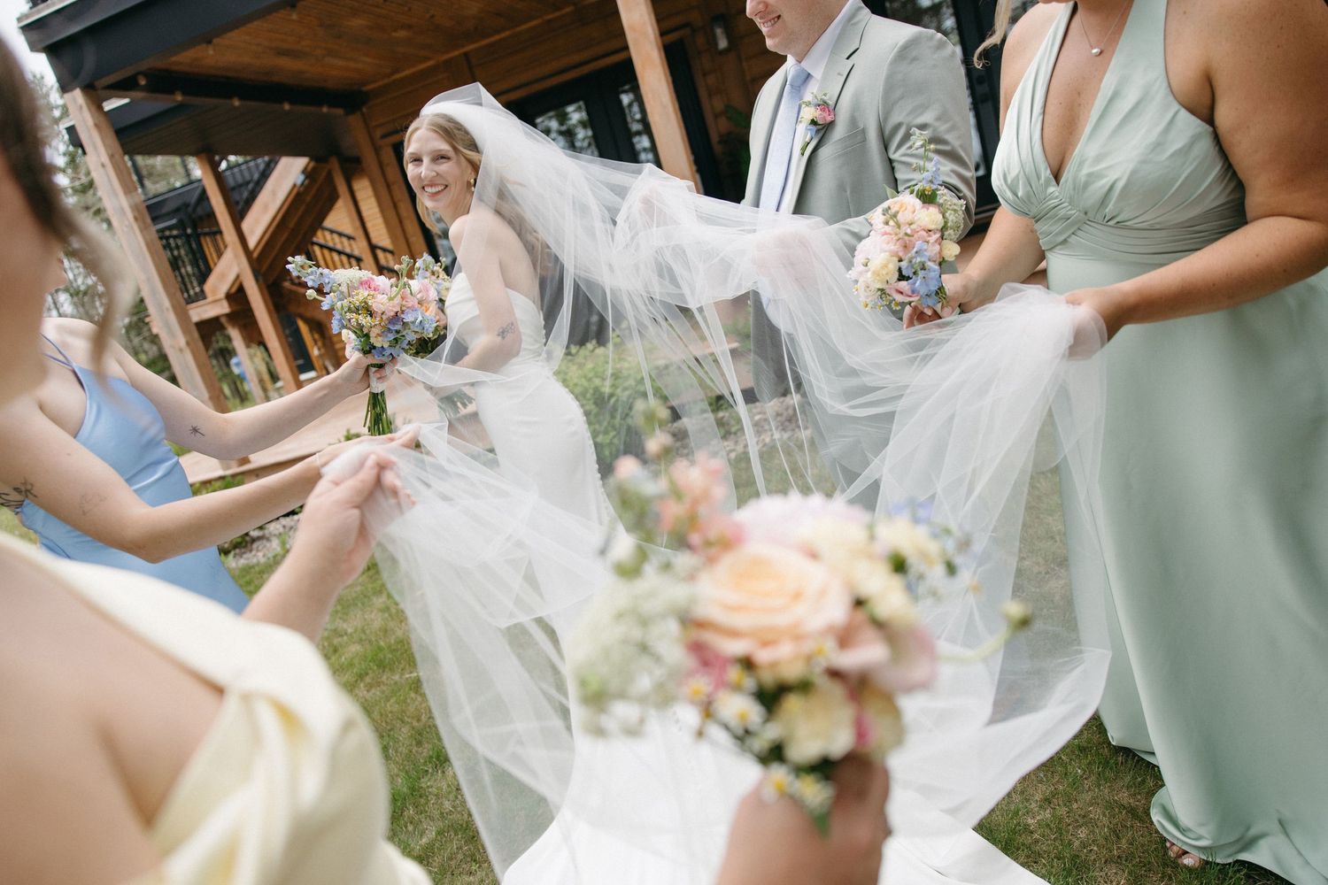 Bridesmaids in pastel gowns holding colorful bouquets while lifting bride's flowing veil at outdoor wedding.