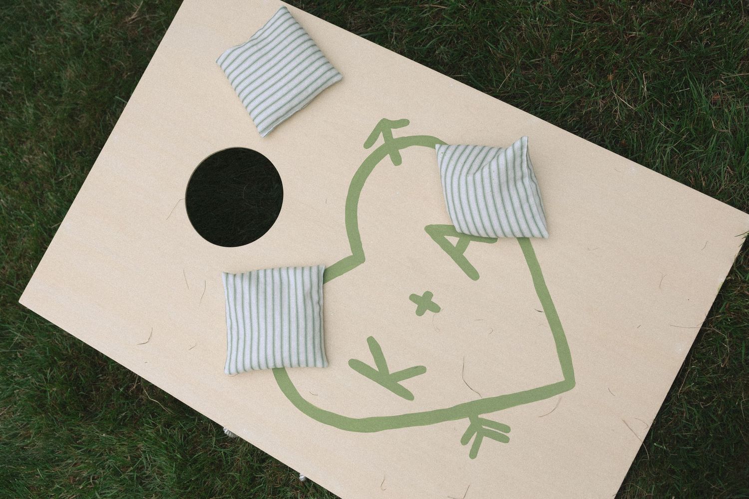 Cornhole board game with frog design and striped bean bags on green grass.